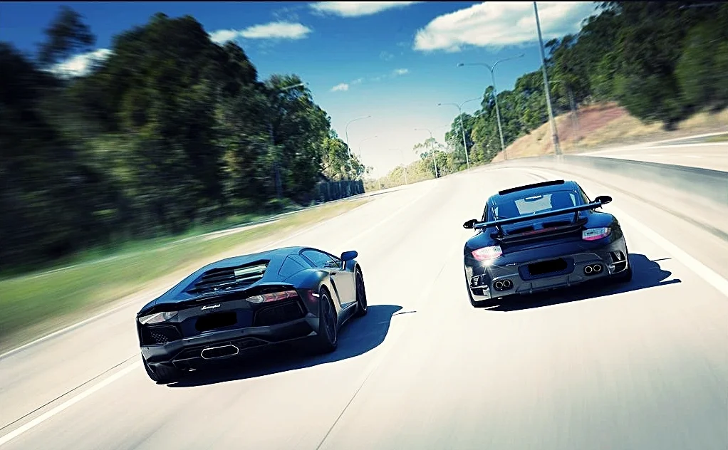 Black Lamborghini Aventador and Porsche 911 driving side by side on a tree-lined motorway