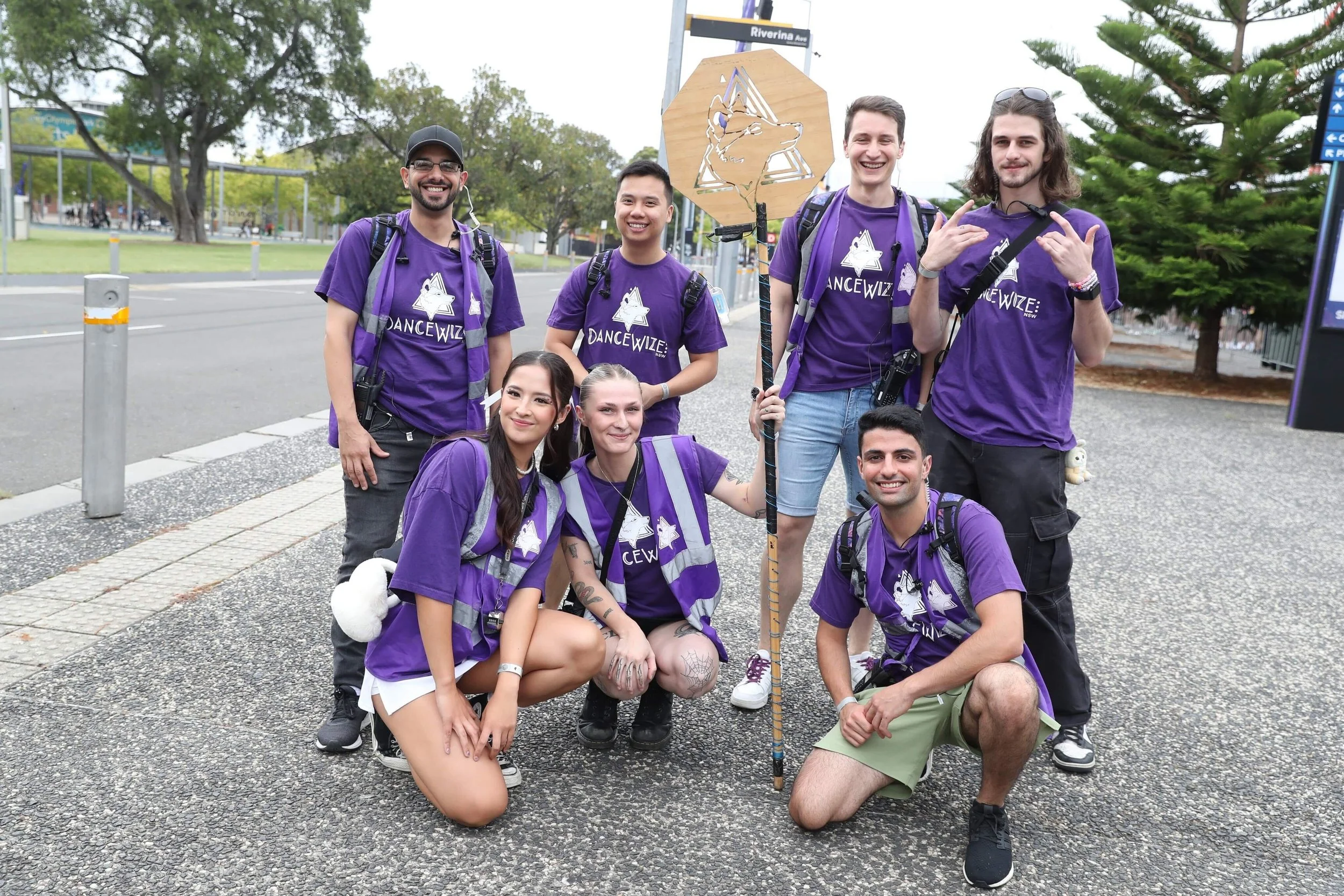 A group of seven people in purple t-shirts poses together, holding a sign and smiling against an outdoor background.