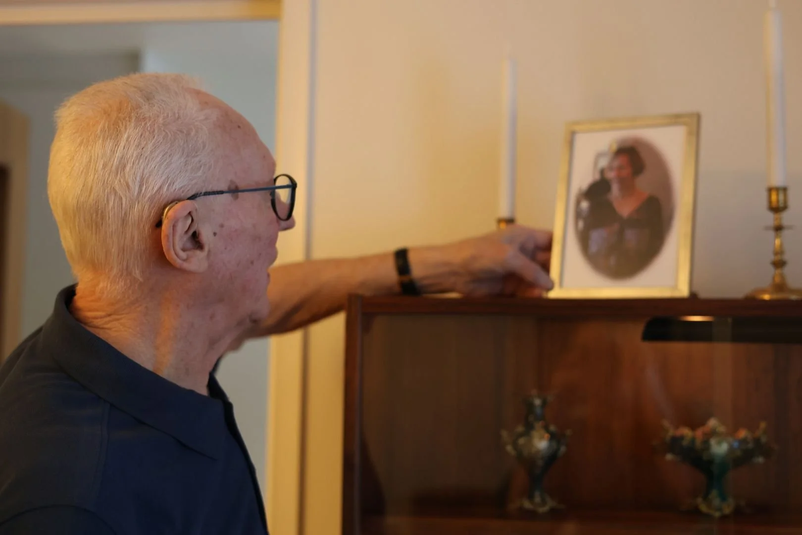 En äldre man i profil med glasögon och hörapparat sträcker sig efter ett inramat fotografi av en kvinna som står på ett mörkt träskåp. Två vita ljus i mässingsstakar flankerar bilden i bakgrunden.
