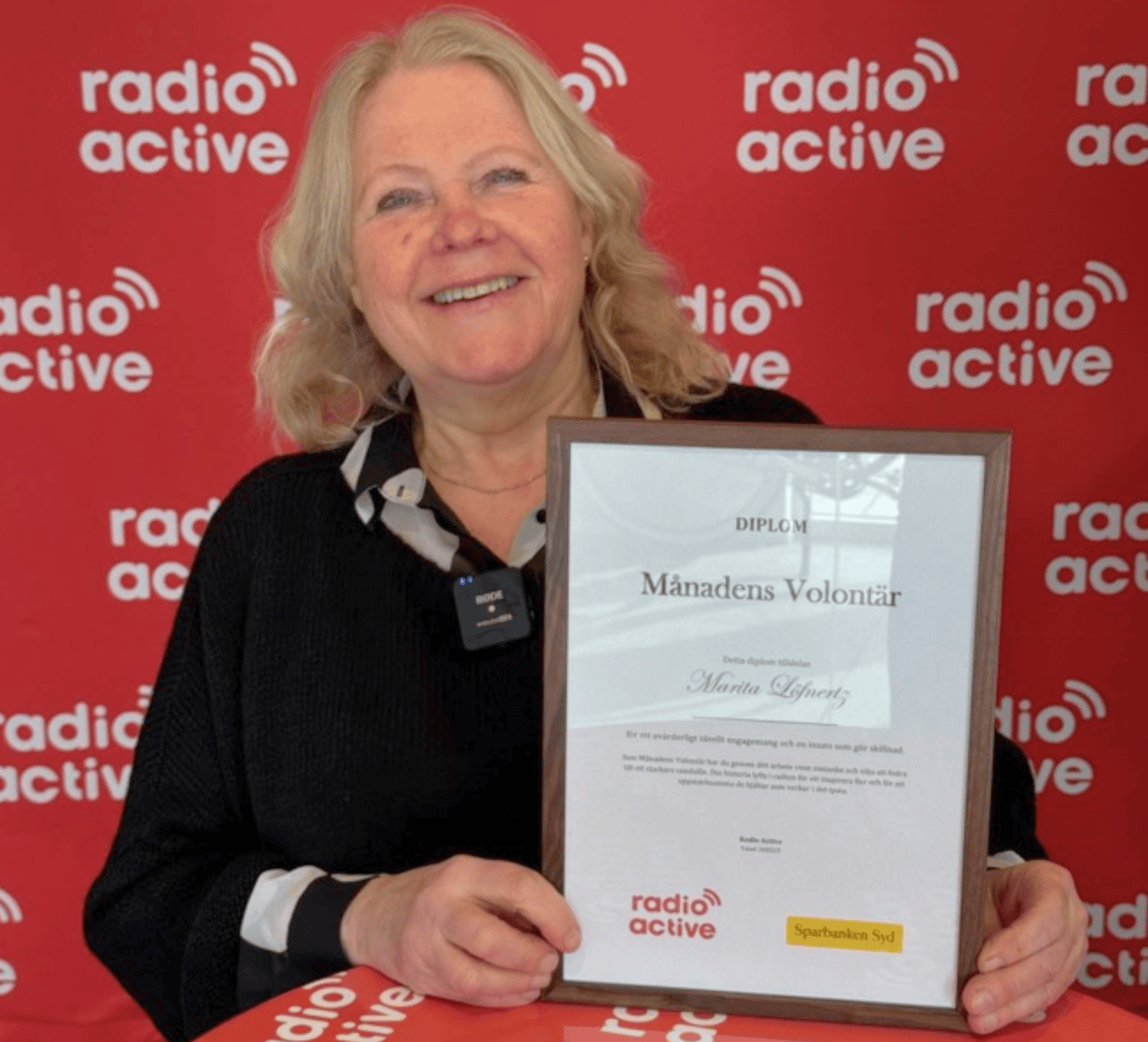 En leende kvinna med ljust hår håller upp ett inramat diplom för ”Månadens Volontär” framför en röd vägg med Radio Actives logotyp.
