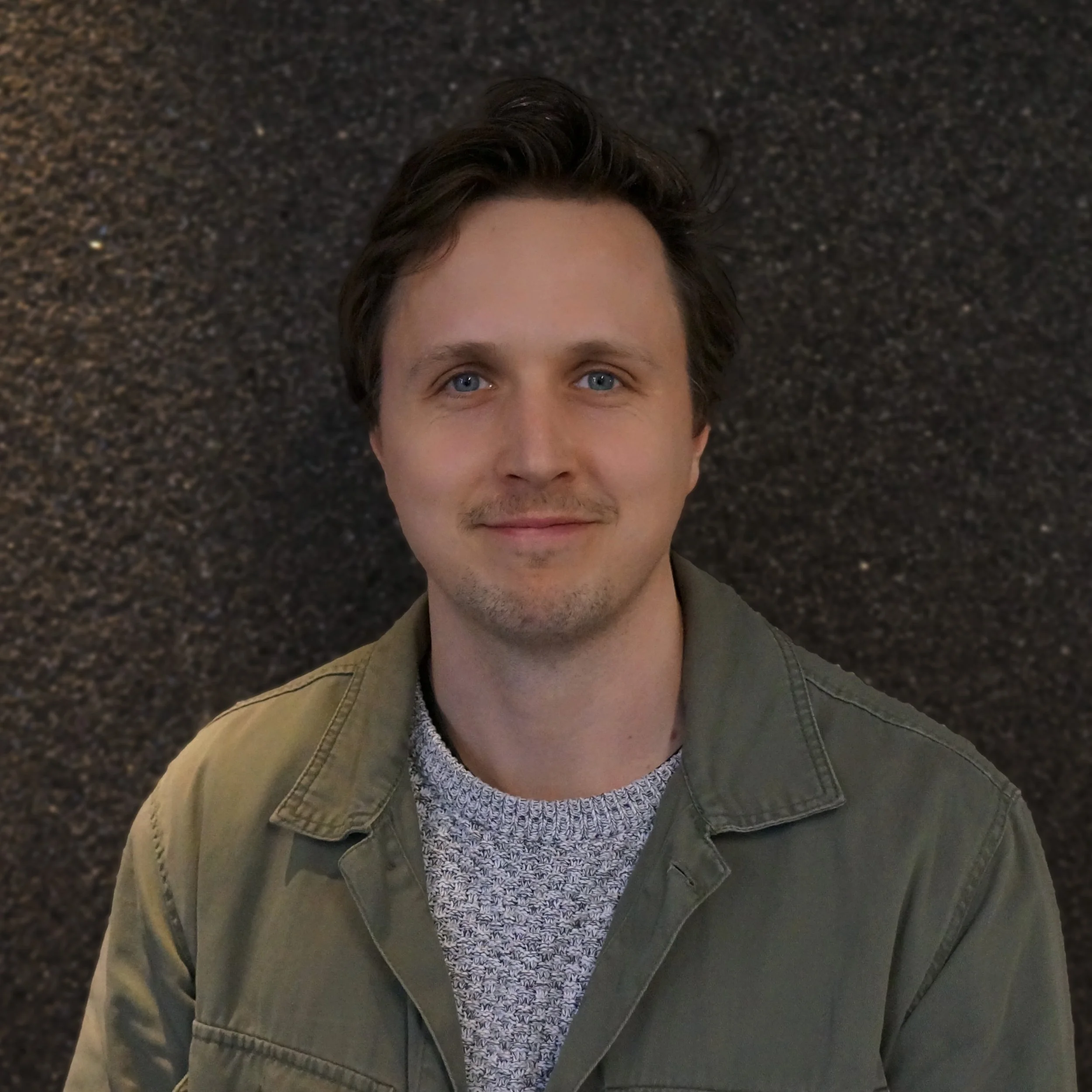 Porträtt av en man med ljus hud, blå ögon och mörkbrunt, lätt vågigt hår, som ler svagt, klädd i en grön jacka och en ljus grå tröja, med en mörk bakgrund.