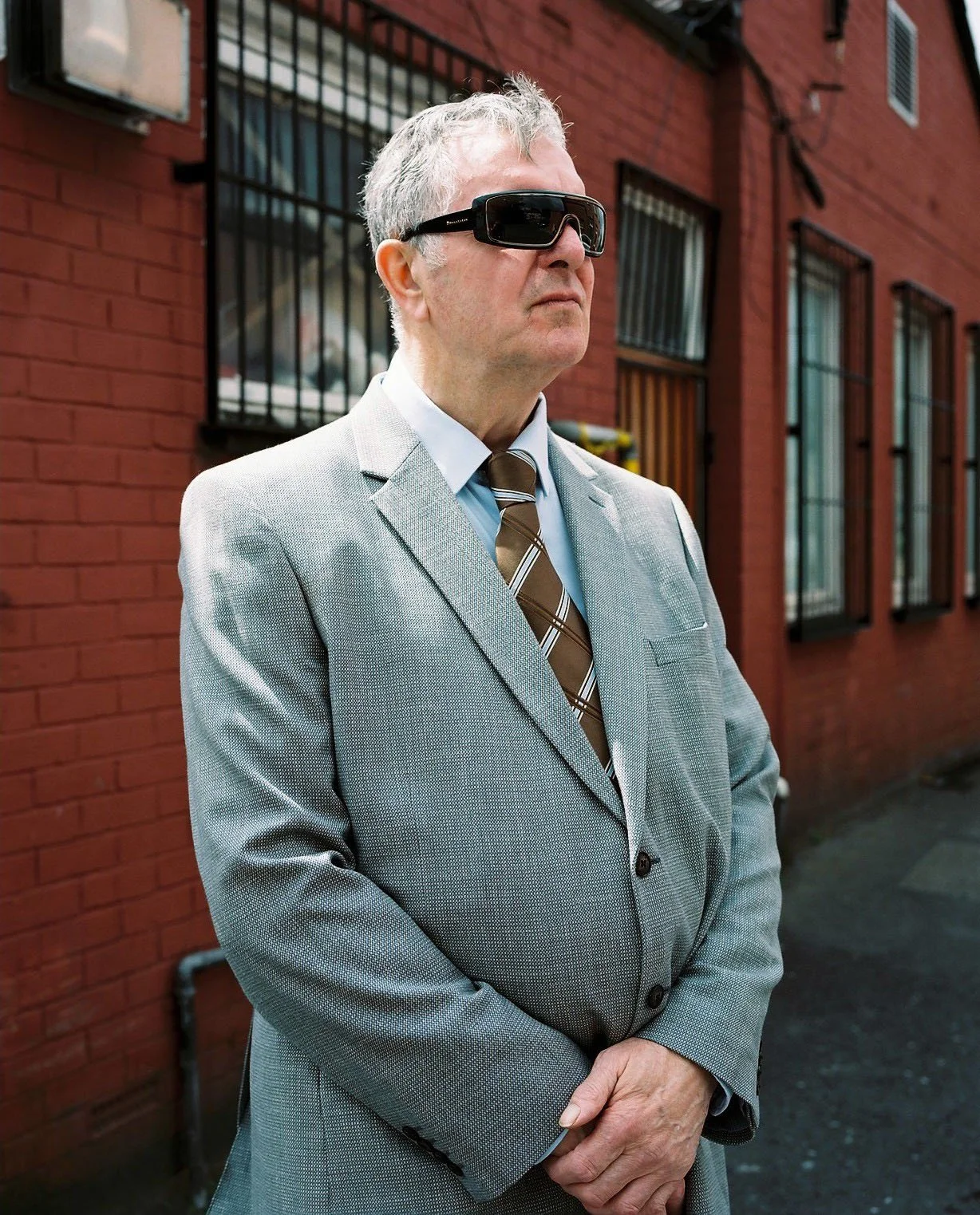 Man with large sunglasses stands outside brick building.