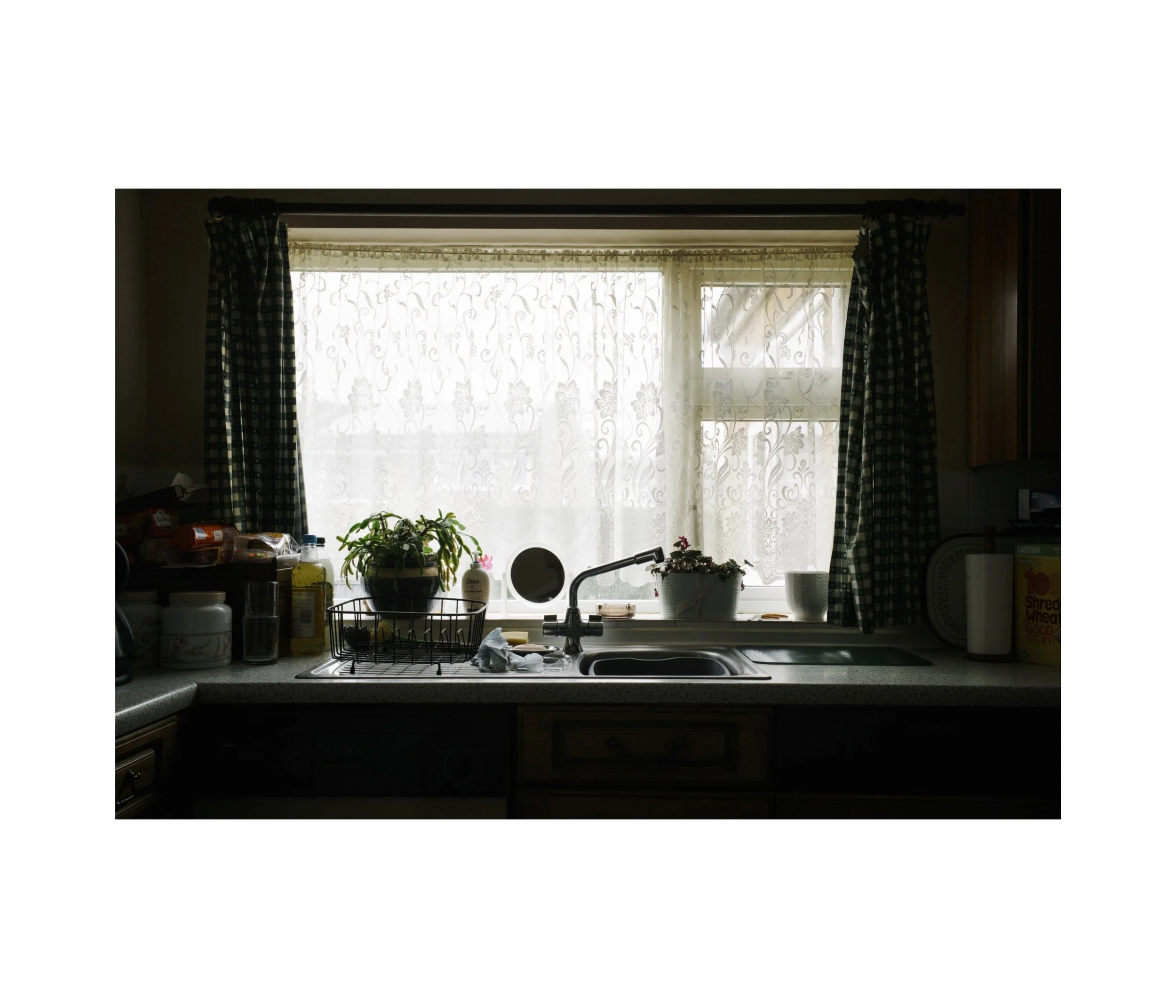 Kitchen sink under a window with net curtains; green check curtains and plants on the sill.