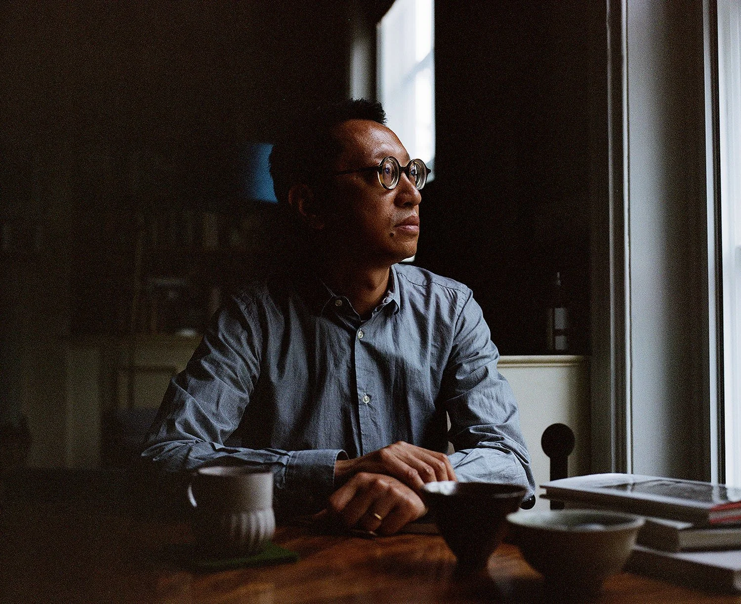Man with round glasses sitting at a table in window light.