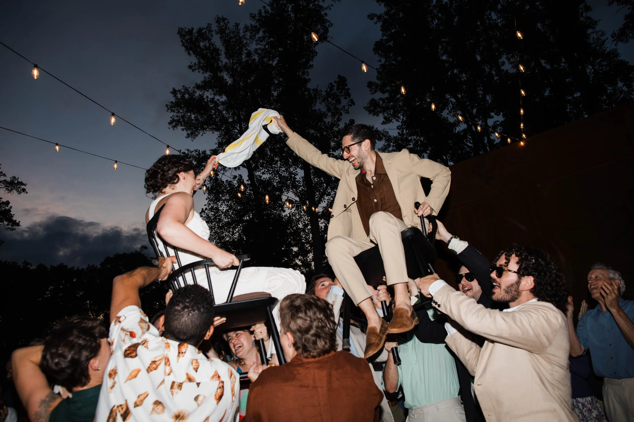 A man is sitting on someone's shoulders during a celebration at night. They are surrounded by a crowd, everyone appears joyful, and the man is reaching out to shake hands with a girl sitting on a chair, all under hanging string lights.