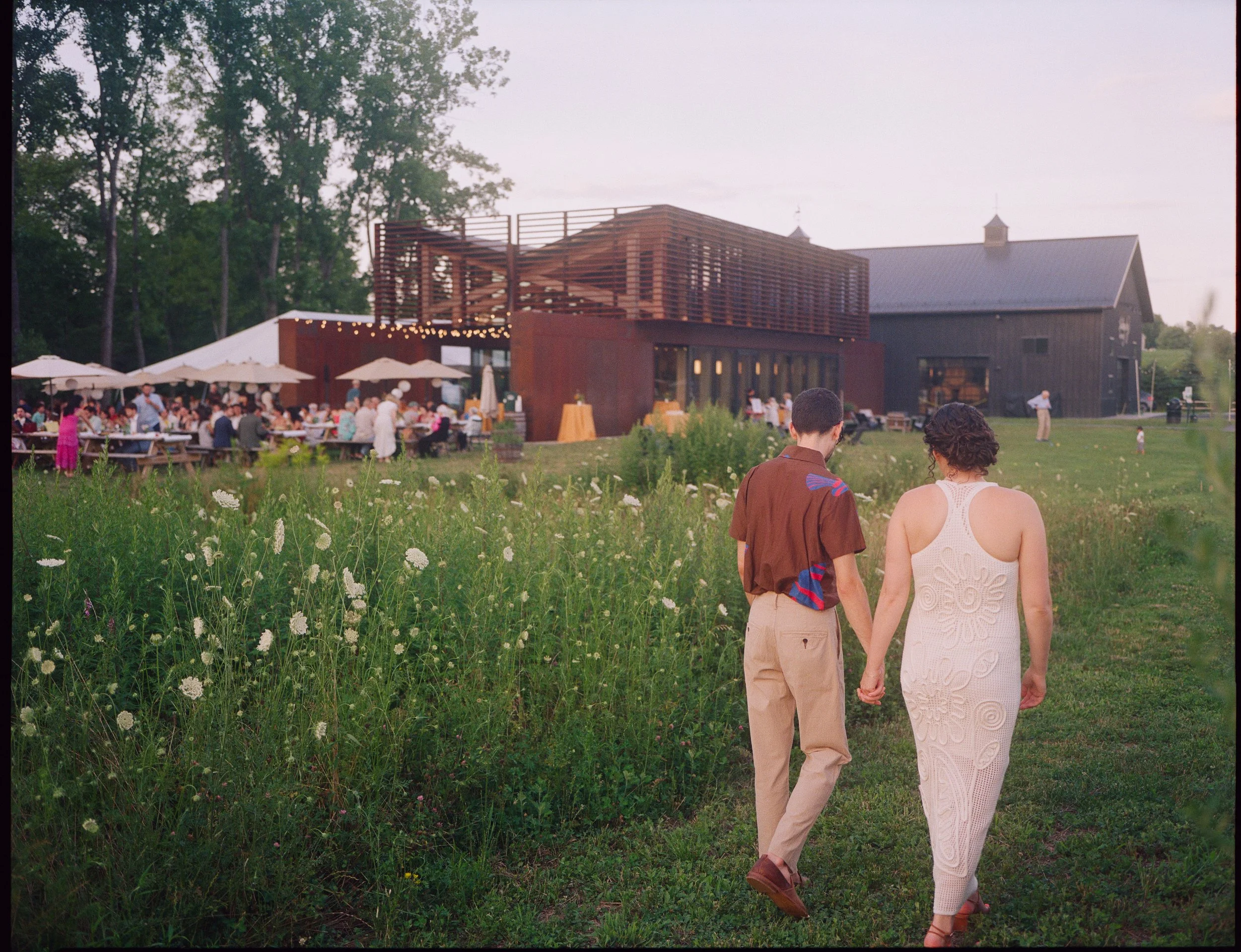 A couple holding hands and walking through a field of white wildflowers towards an outdoor event with tables, umbrellas, and a large wooden building in the background, with trees and a cloudy sky overhead.