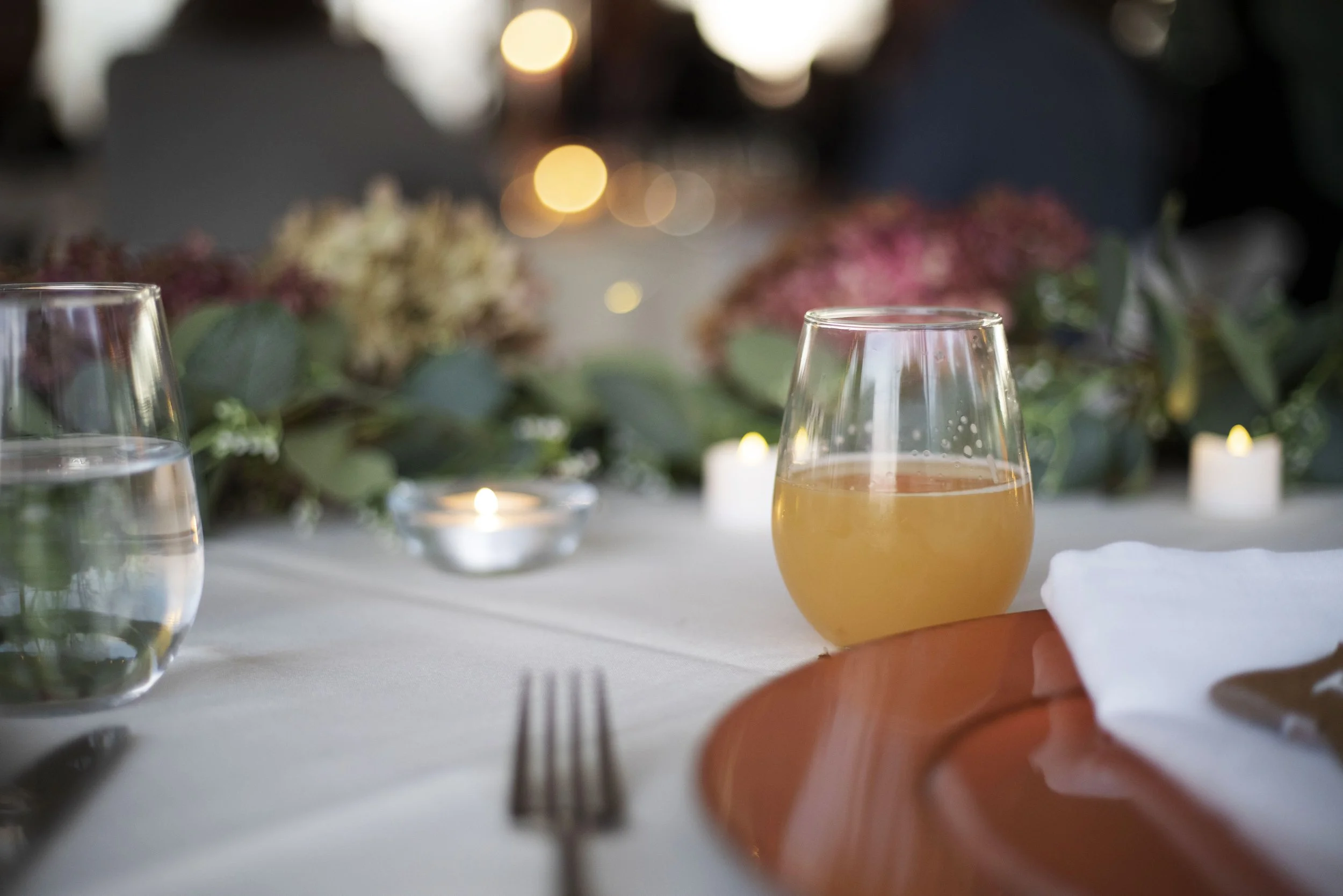 Close-up of a wine glass and a beer glass on a table with blurred flowers and candles in the background.