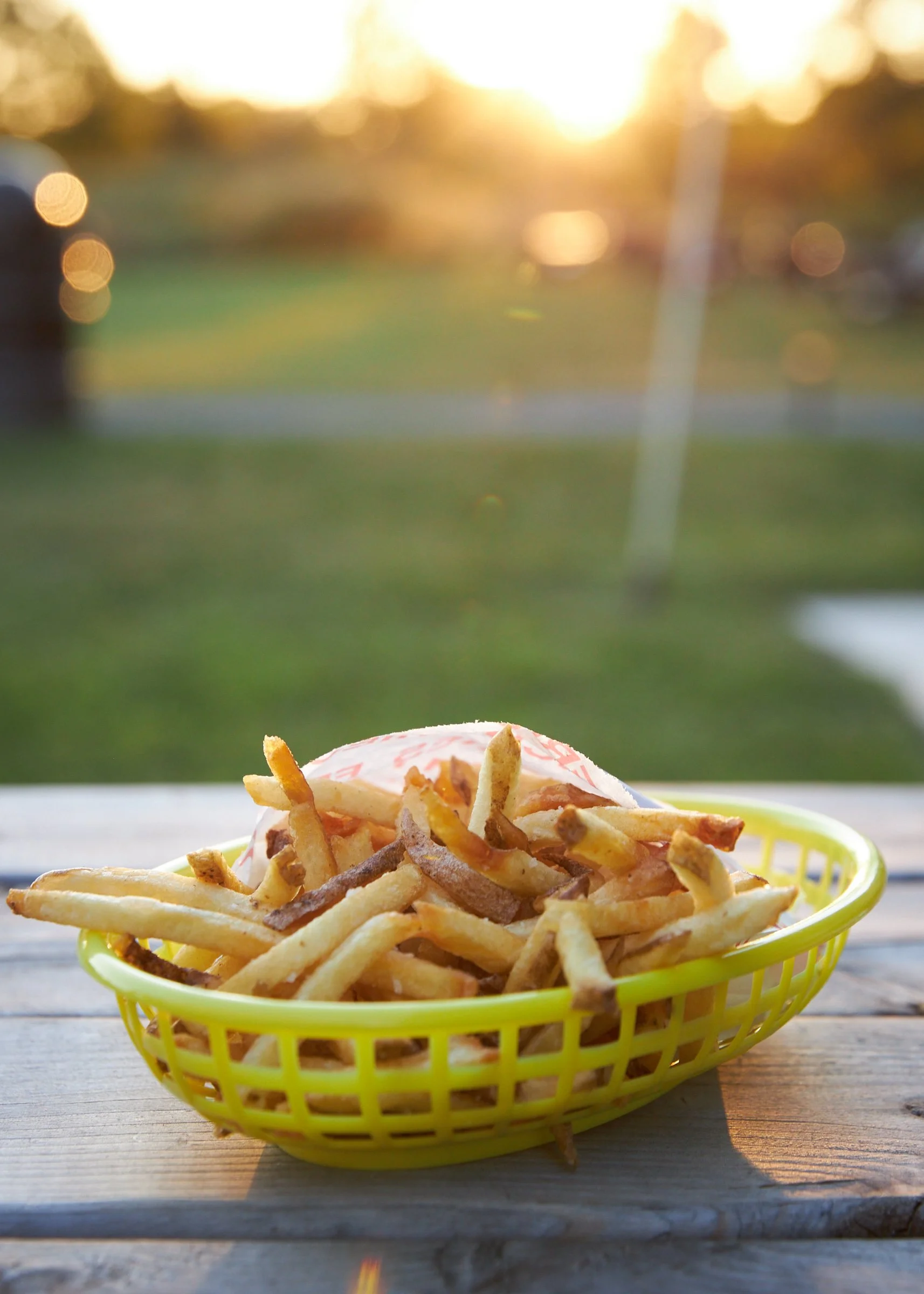 Basket of French fries on a wooden table with a blurred outdoor background.