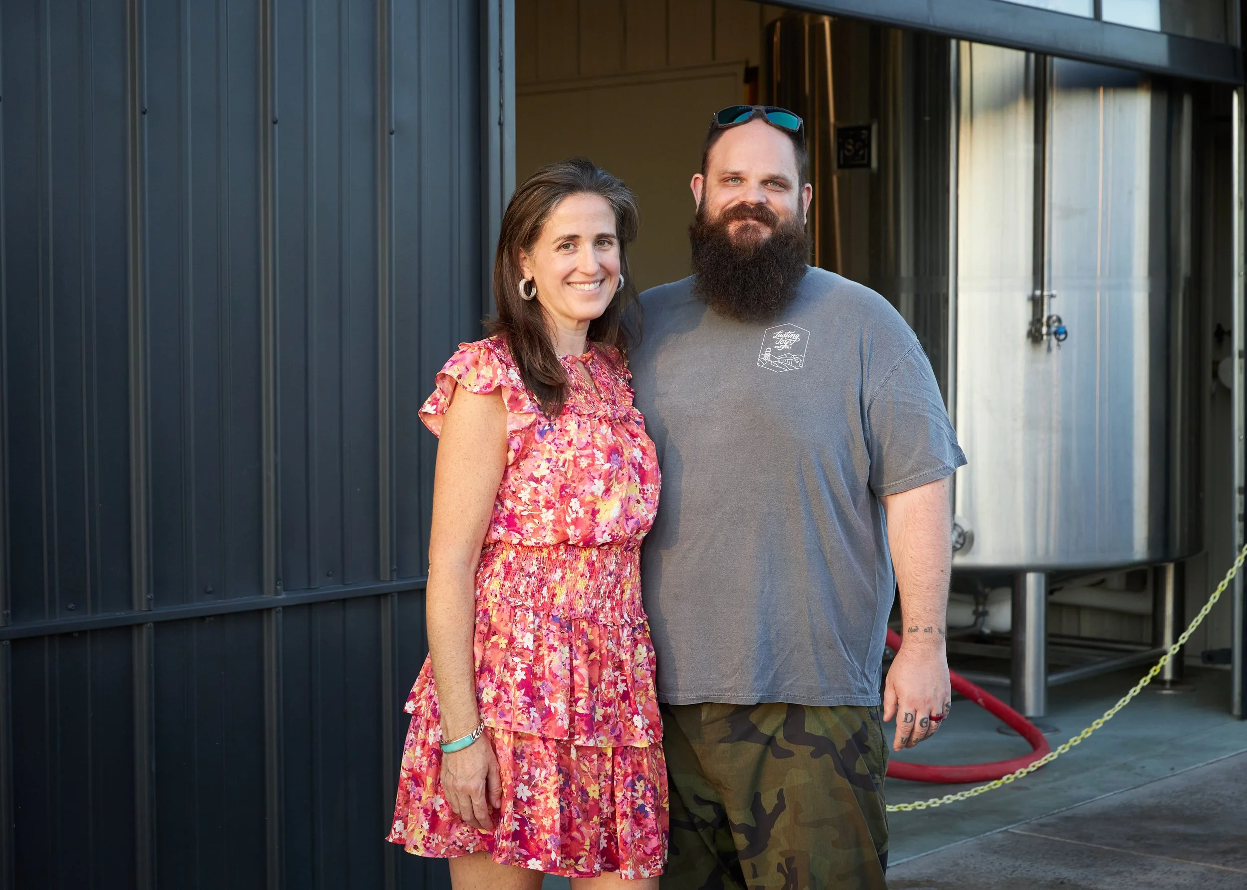 A smiling woman with long brown hair wearing a pink floral dress and large earrings standing next to a bearded man with short hair wearing sunglasses, a gray T-shirt, and camouflage shorts, outdoors near a black building with a chain barrier.