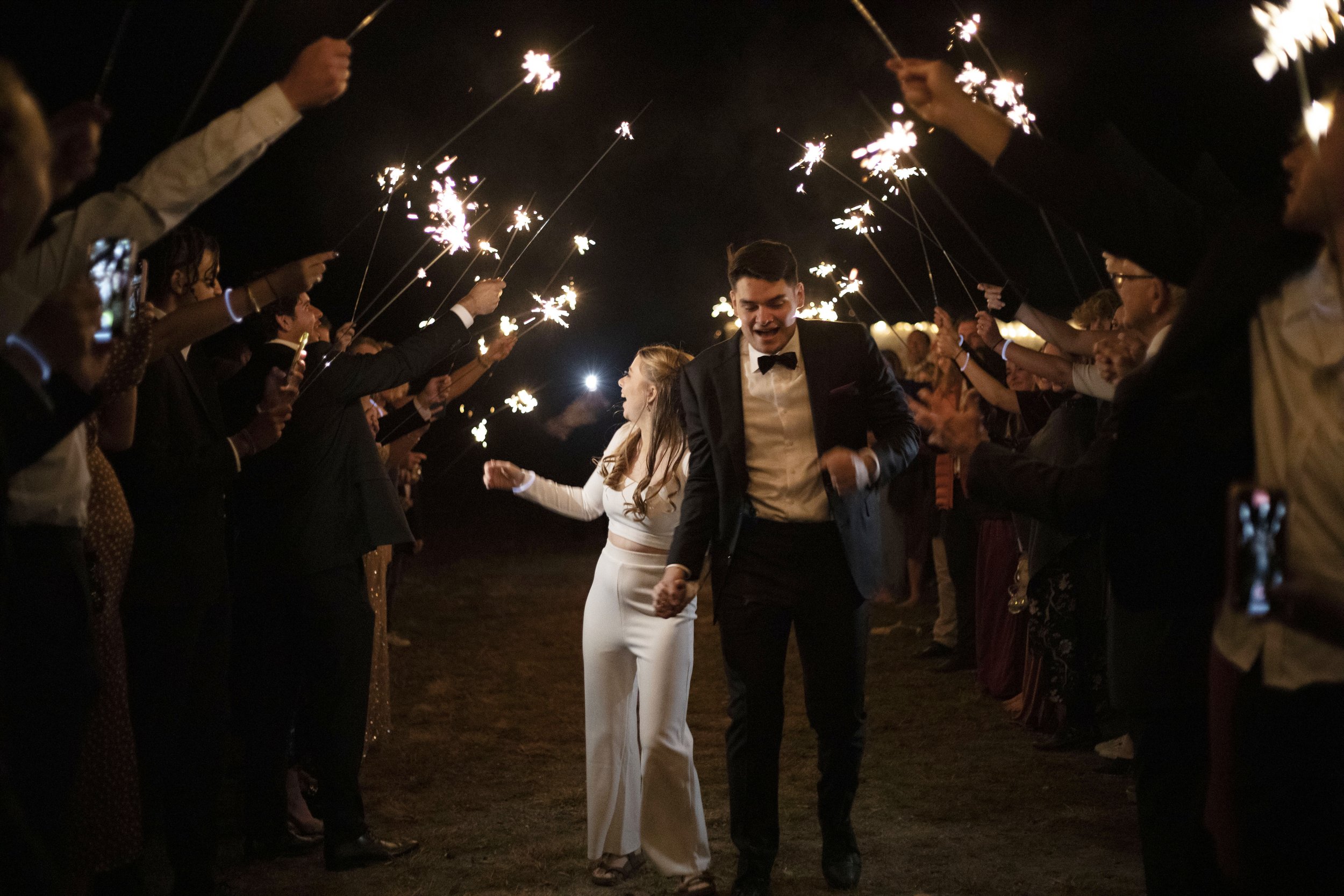 A couple dressed in formal wedding attire walking hand in hand through a crowd holding sparklers at night.