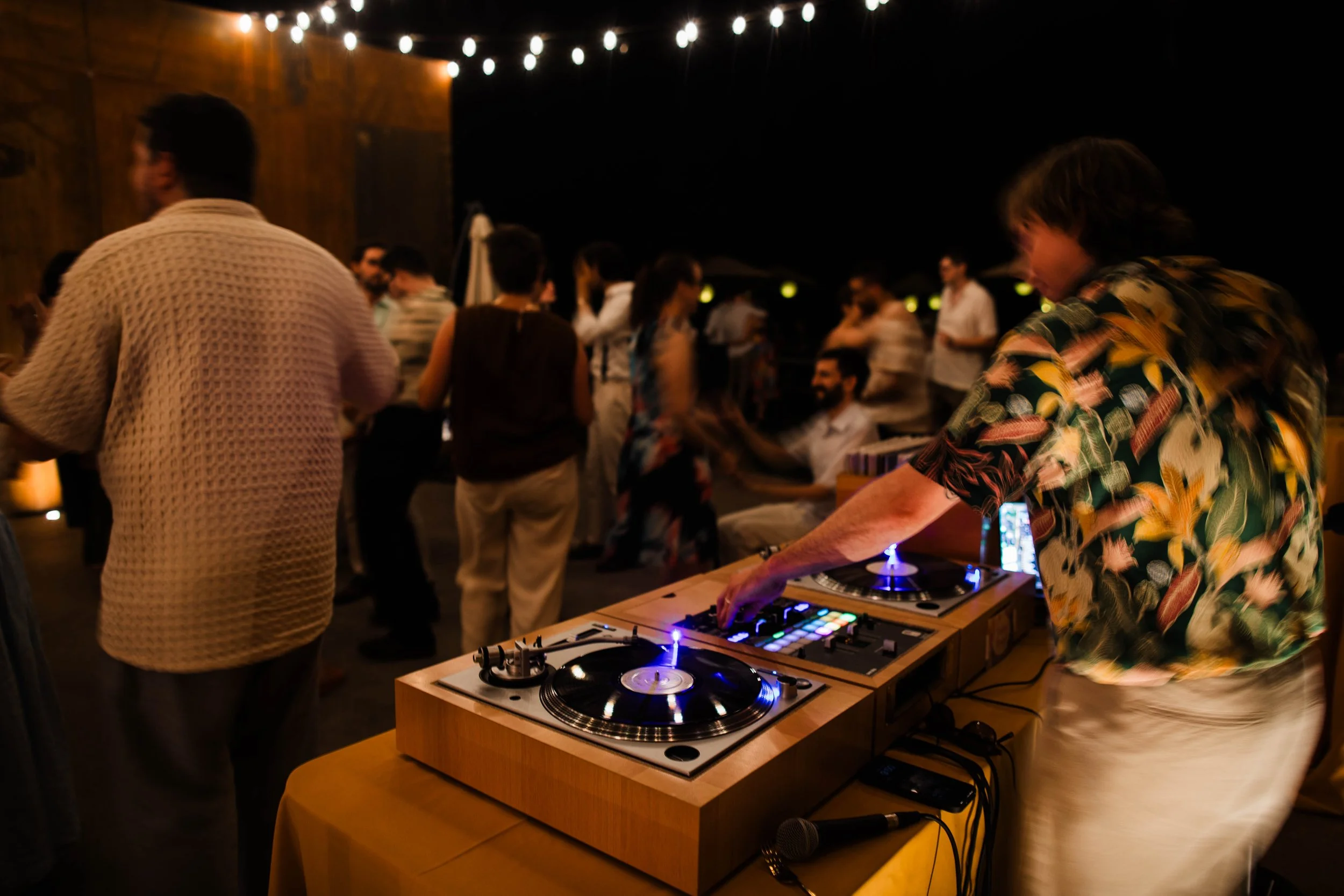 A vinyl DJ spins records under twinkling patio lights as guests dance and enjoy a party on a patio on a warm night. 