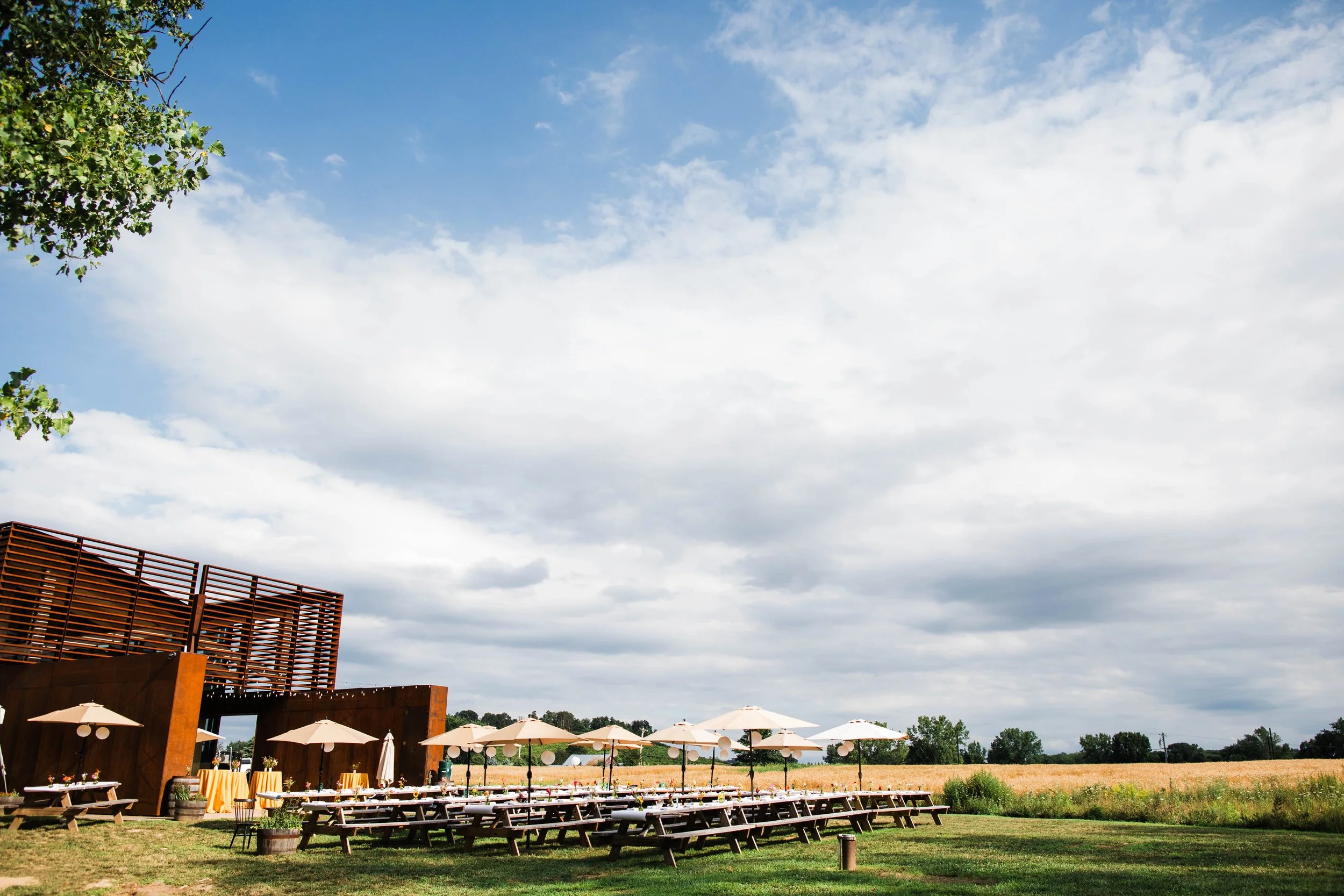 Outdoor event space with rows of picnic tables and umbrellas, a wooden building with slatted walls, grassy field, and partly cloudy sky.
