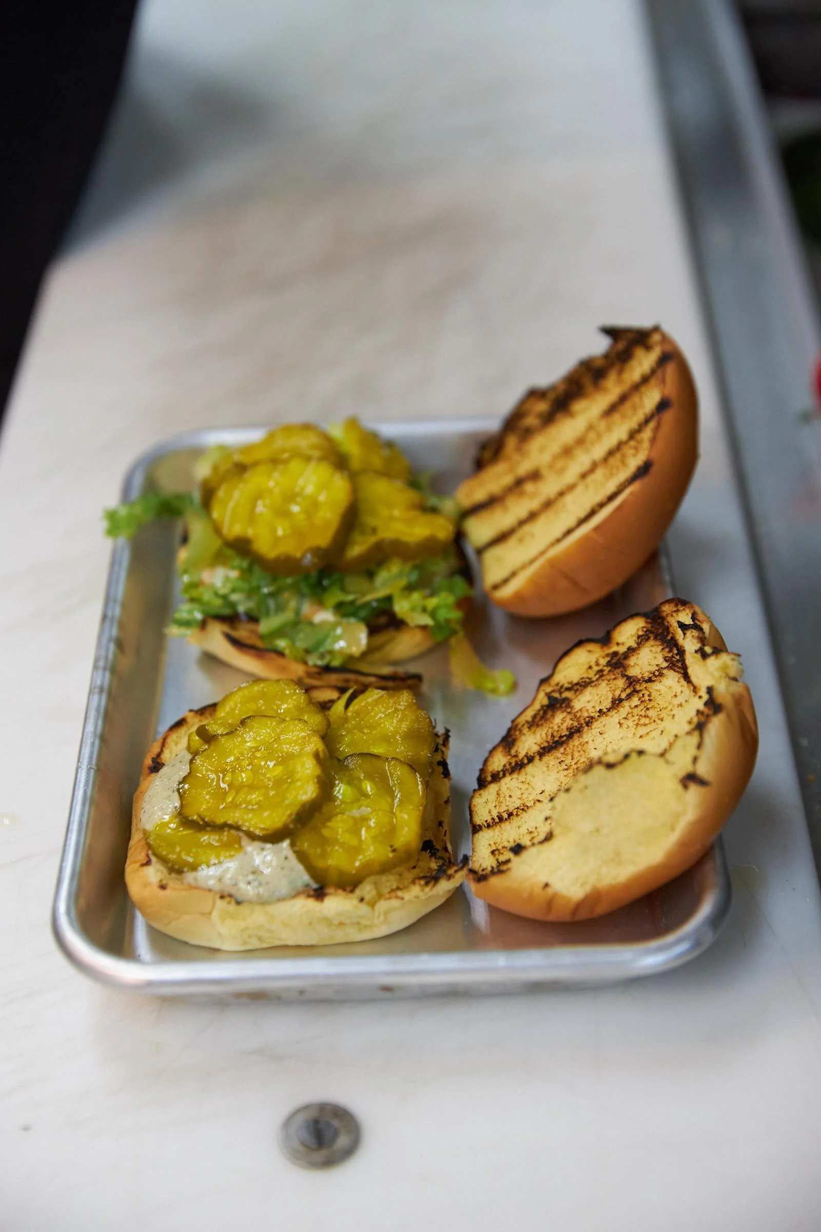Close-up of a metal tray with three grilled burger buns topped with sliced pickles. The sandwiches are on a white table.