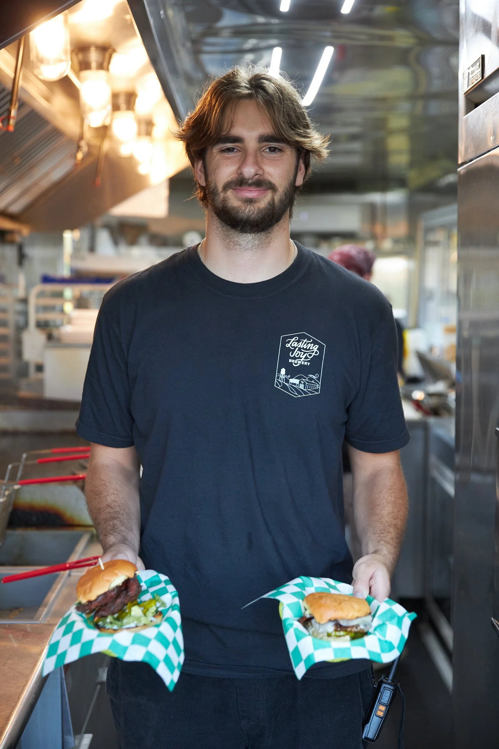 A young man with a beard and wearing a black T-shirt holding two burgers wrapped in green and white checkered paper in a commercial kitchen.