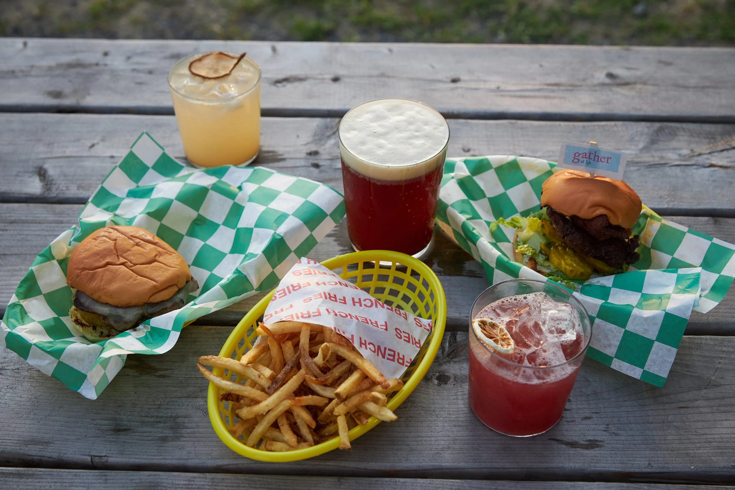 Two burgers, a side of fries, and three glasses of drinks on a wooden outdoor table, with a grassy background.