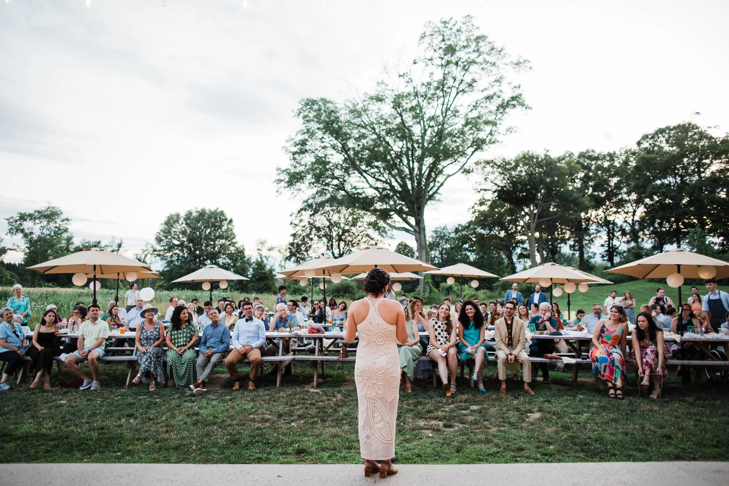 A woman giving a speech to an outdoor audience seated on picnic tables, with large umbrellas, in a park with tall trees in the background.