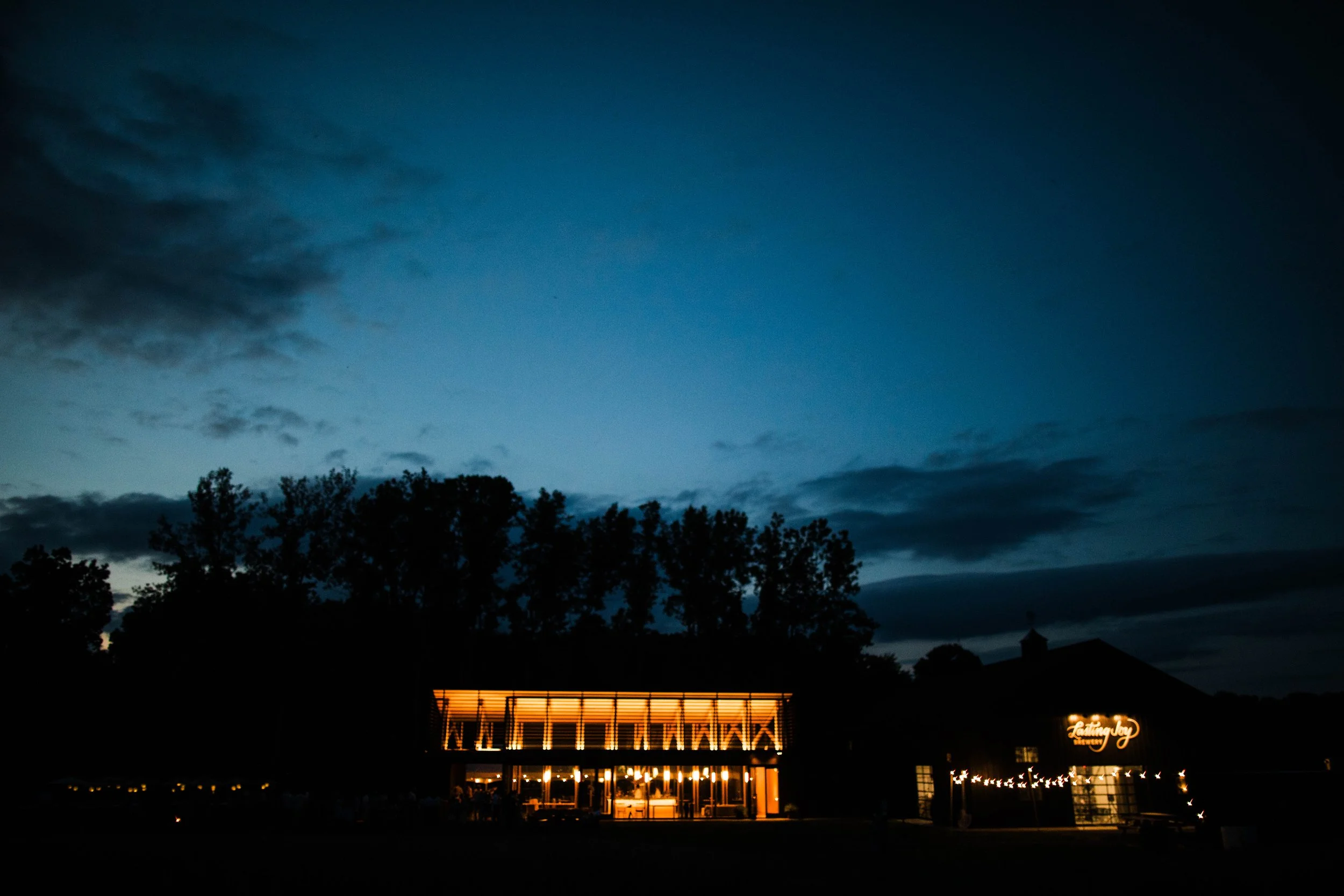 A building illuminated with warm orange and yellow lights at night, with a sign that says "Loving Joy" and string lights outside. The sky is dark with some clouds and silhouettes of trees in the foreground.