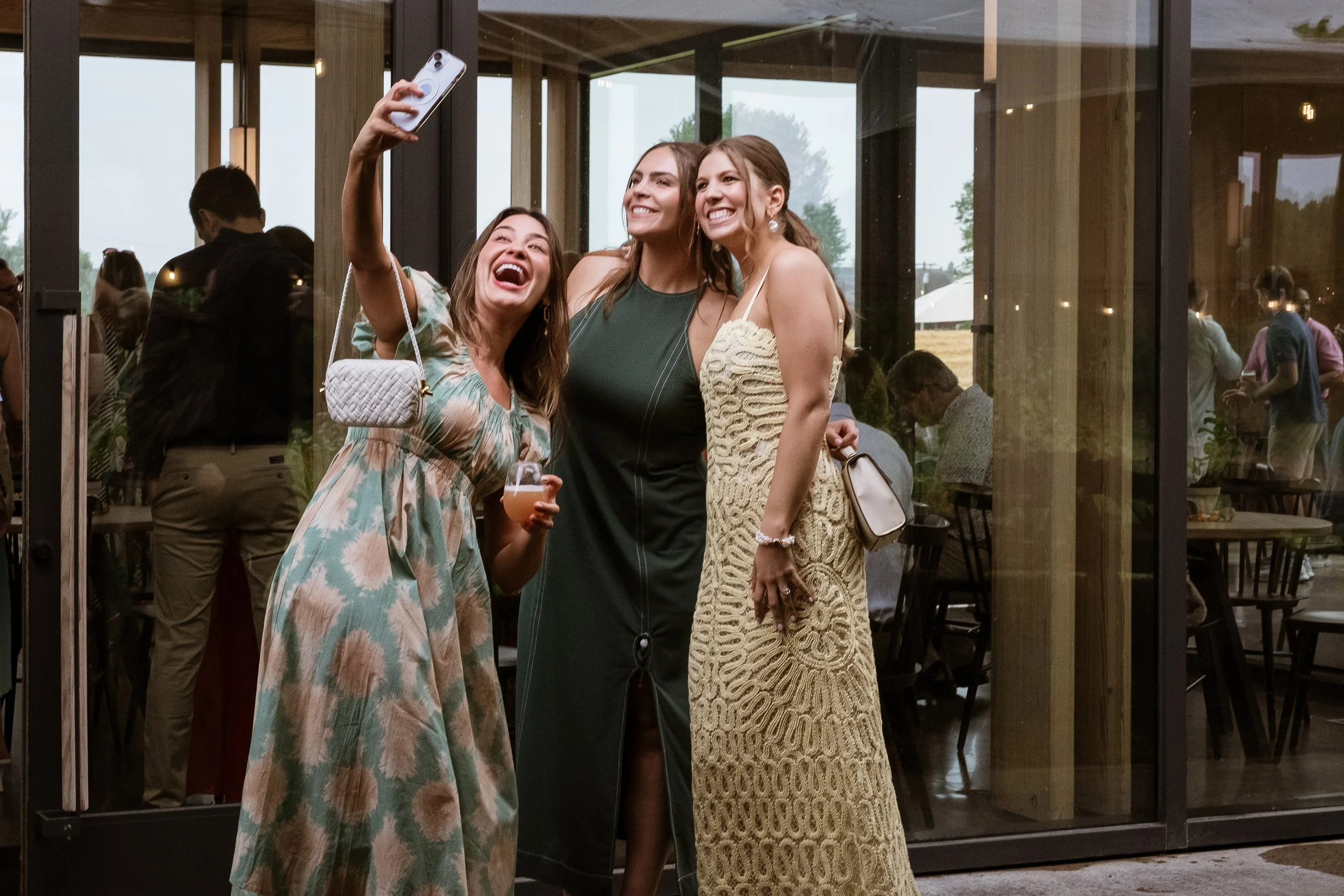 Three women in long dresses at a summer time party pause to take a smiling selfie together while the joyful party is taking place in the background. 
