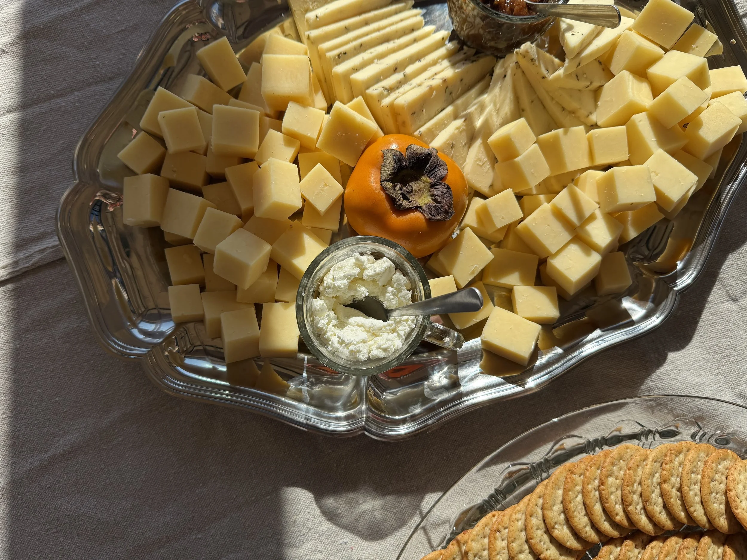 A silver serving tray filled with various types of cheese cut into cubes, including blue cheese and cheddar, along with a small dish of crumbled cheese, a persimmon fruit, and a spoon. To the right, there are round crackers on a separate dish.