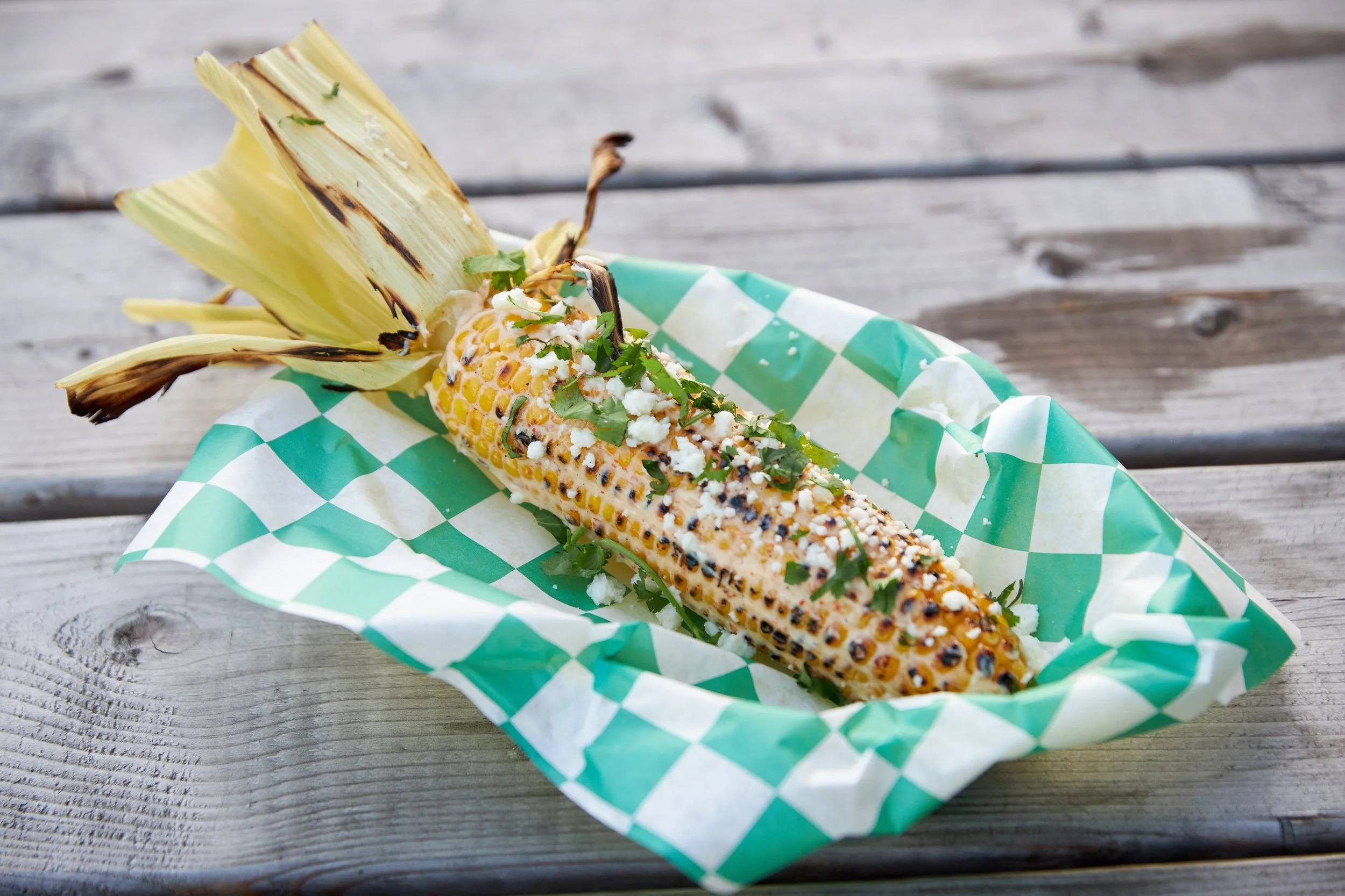 Grilled corn on the cob garnished with chopped herbs and crumbled cheese, served on green and white checkered paper on a wooden surface.