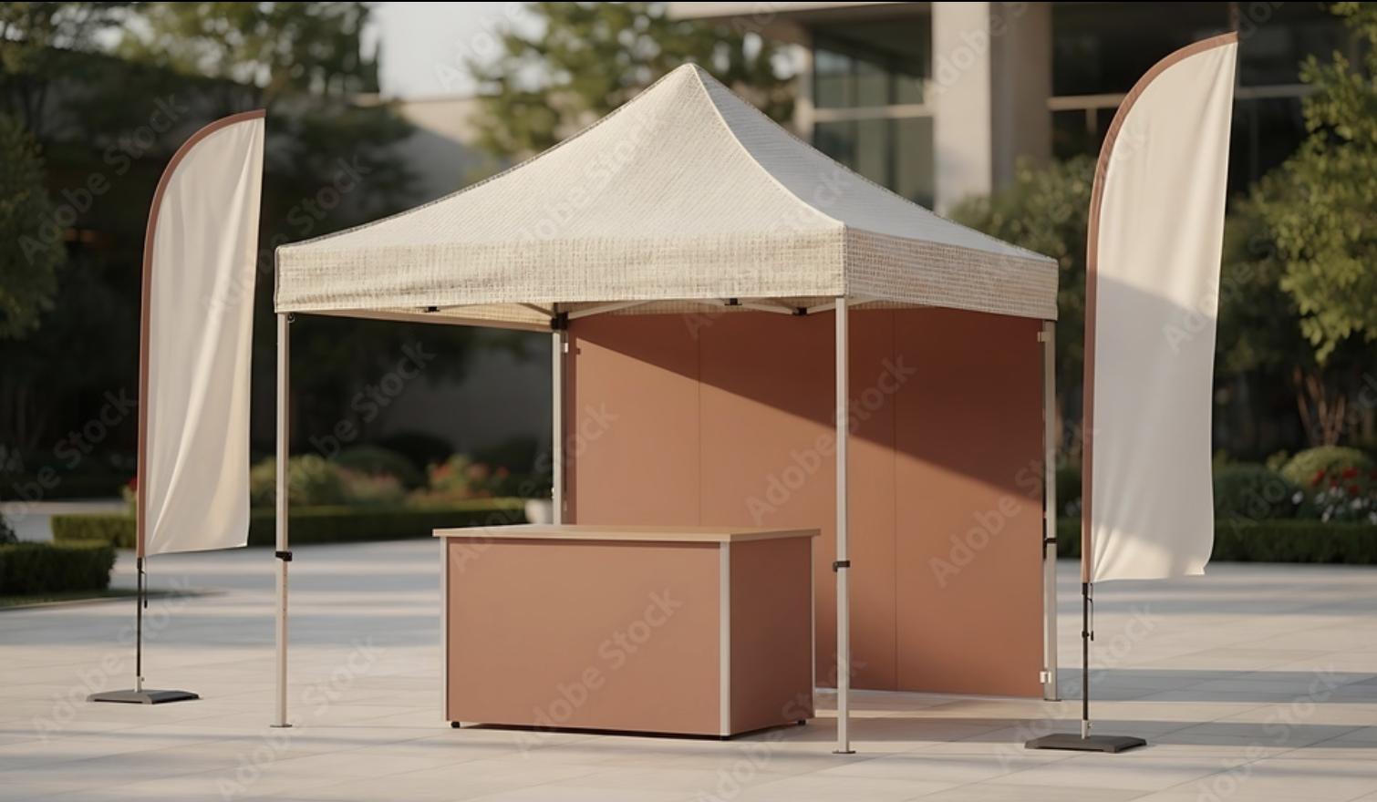 An outdoor booth with a beige canopy, pink background panels, and a pink counter, flanked by two white flags with brown trim, set on a paved area with trees and building in the background.