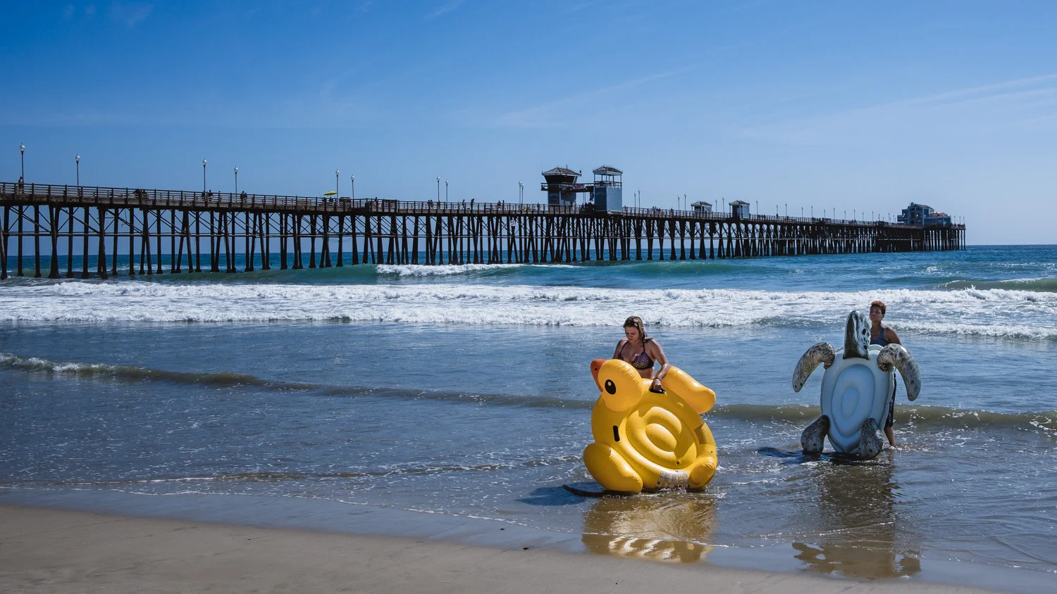 Two girls carry their float toys on the Oceanside beach, San Diego - travel photography and guide by © Natasha Lequepeys for "And Then I Met Yoko". #sandiego #california #travelguide #photoblog #travelblog #usa #sealions #fujifilm #travelphotography #landscapephotography #naturephotography #travelitinerary #2dayssandiego #pwc #westcoast #nature #californiadreaming #pier #oceanside #beach