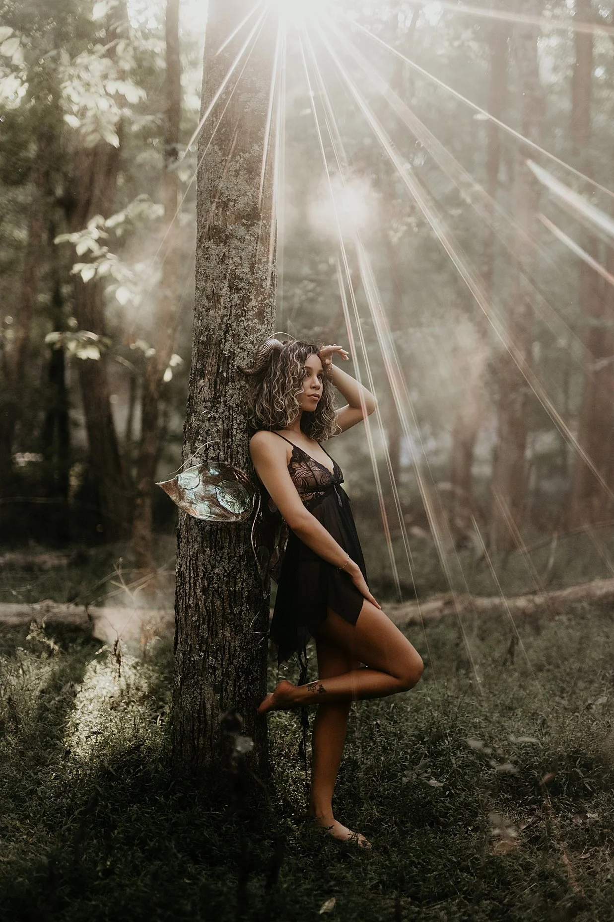 Artistic fantasy photography session featuring a woman standing in a forest beside a tree.
