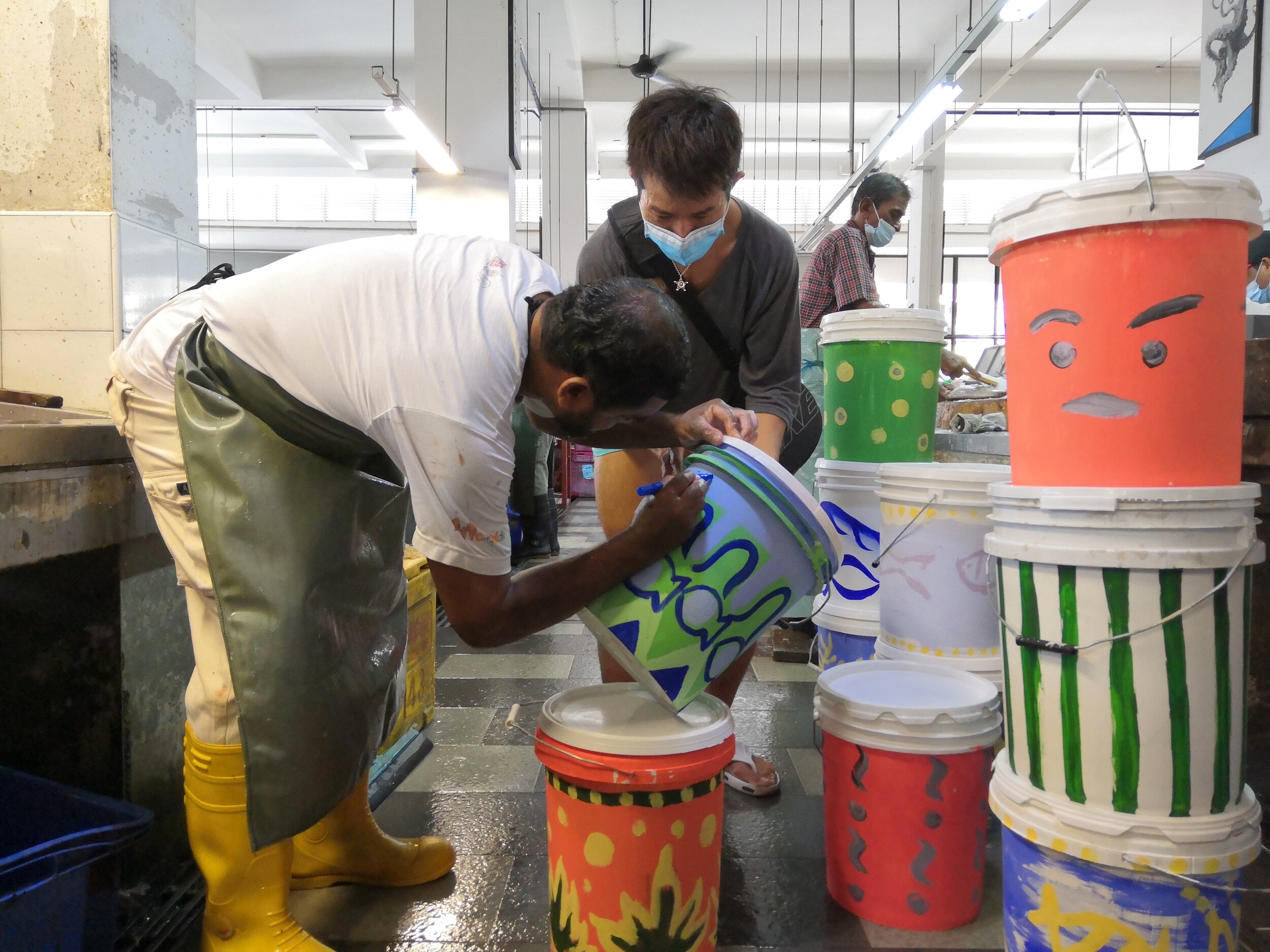  The trainees painted pails which were then distributed to all vendors. One of the vendors, Yasin (left) writing his stall number on his preferred pail. 