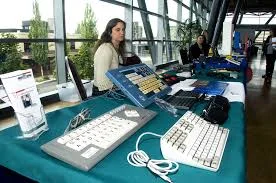 A display of multiple keyboards. One has multiple colored keys, one has large keys, one is slanted. A woman stands behind the table