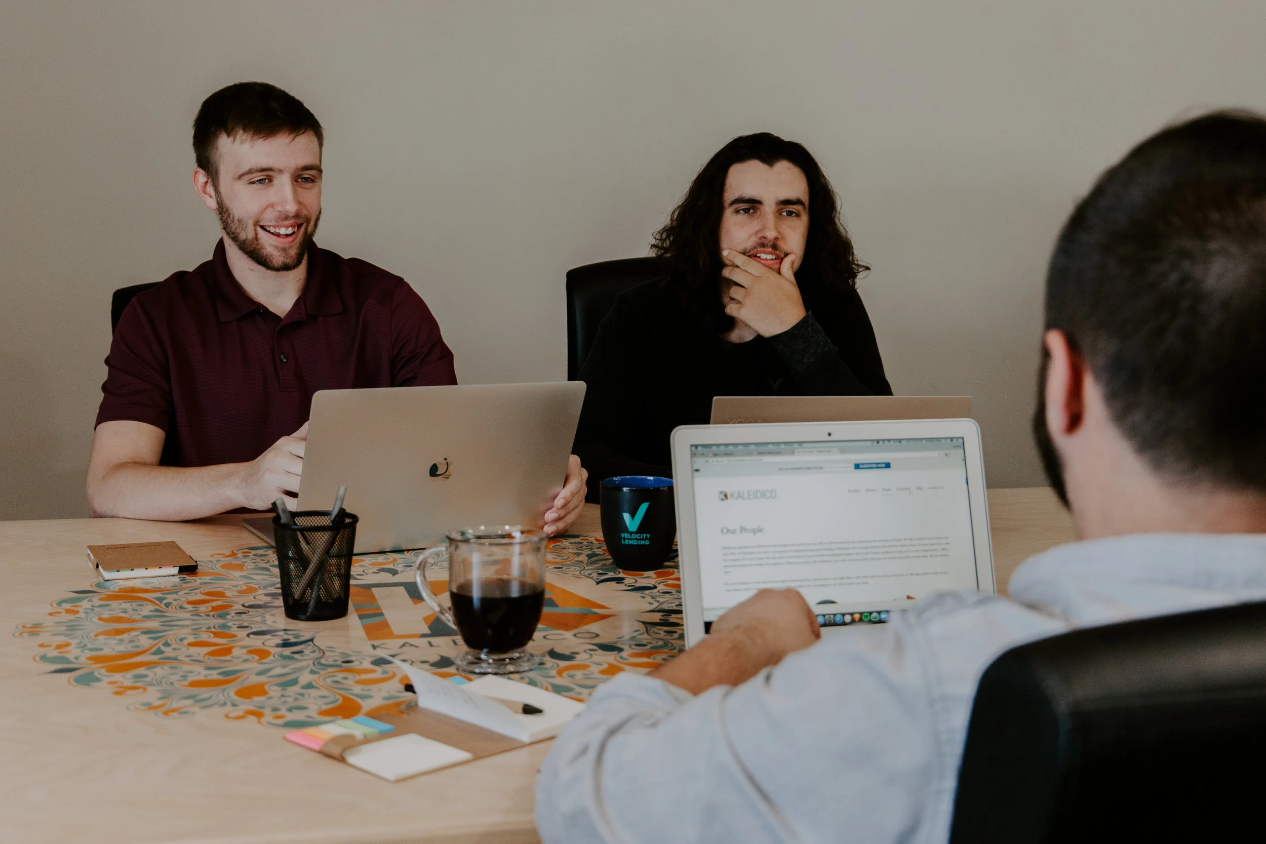 three men sitting around a table with laptops and coffee