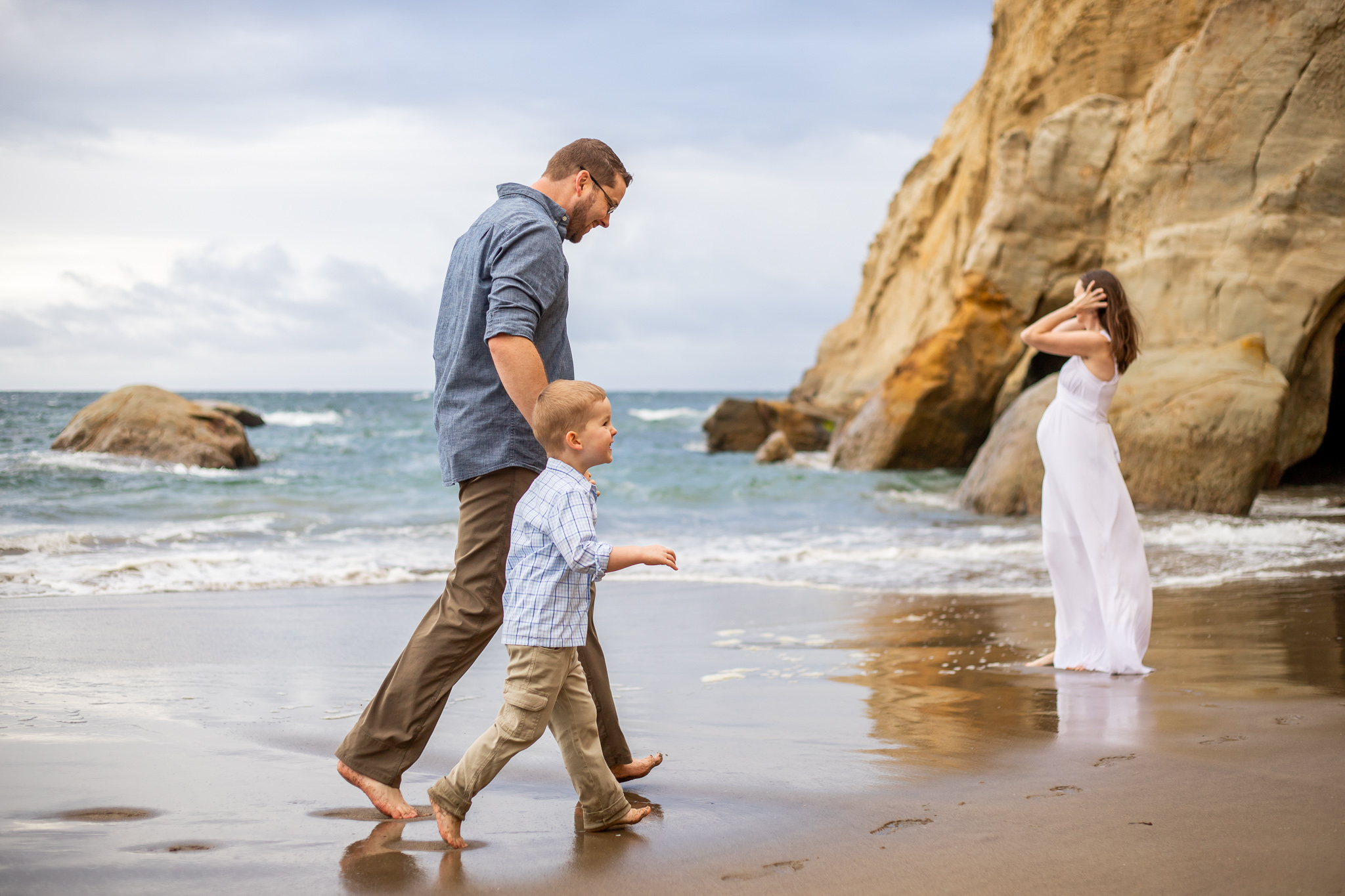 Father and son walk on beach with pregnant mother in a white dress standing on the shore of the ocean.