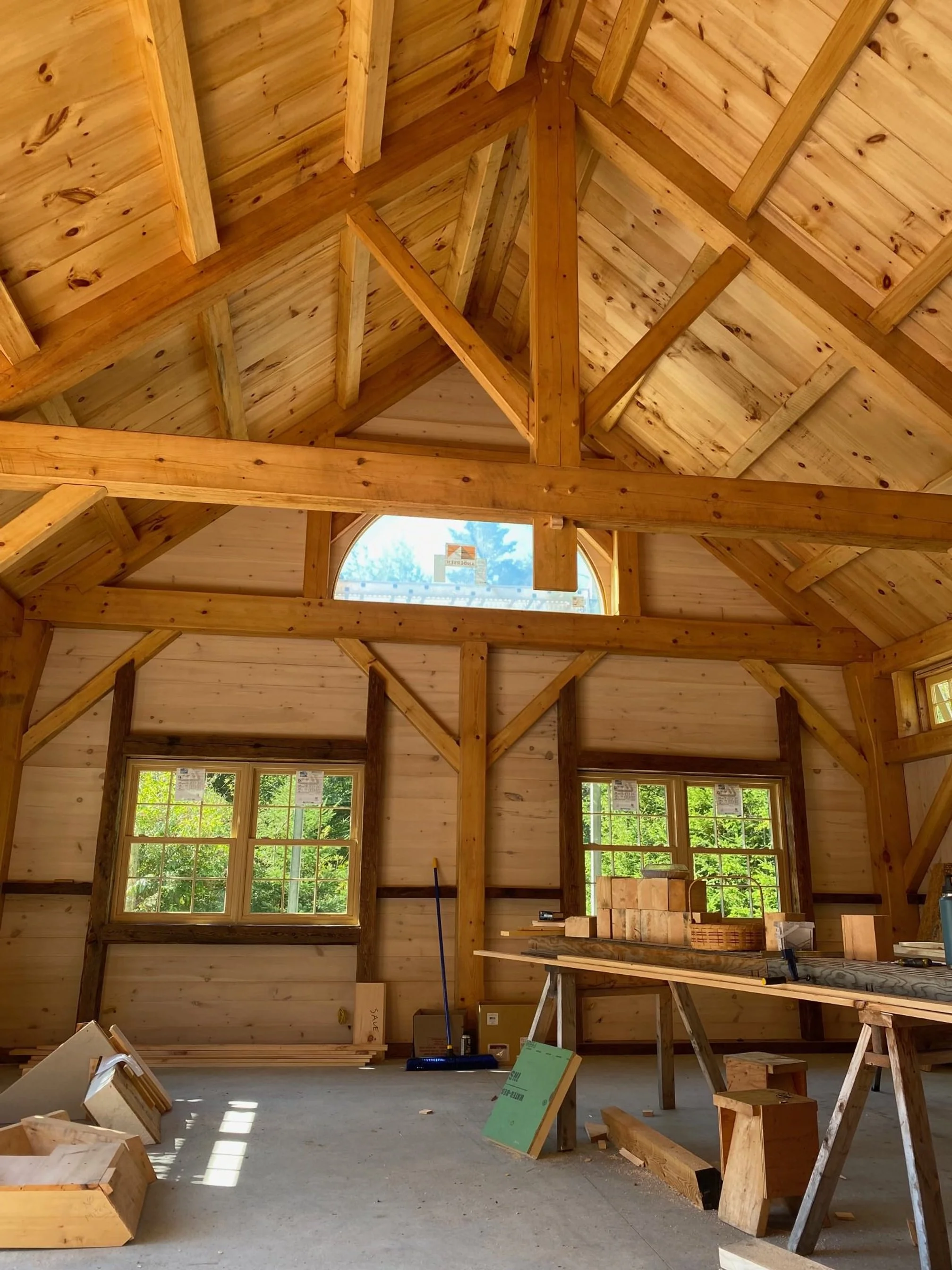 Interior view of a wooden frame house under construction, showing exposed wood beams, plywood walls and ceiling, and a concrete floor with construction materials and tools.