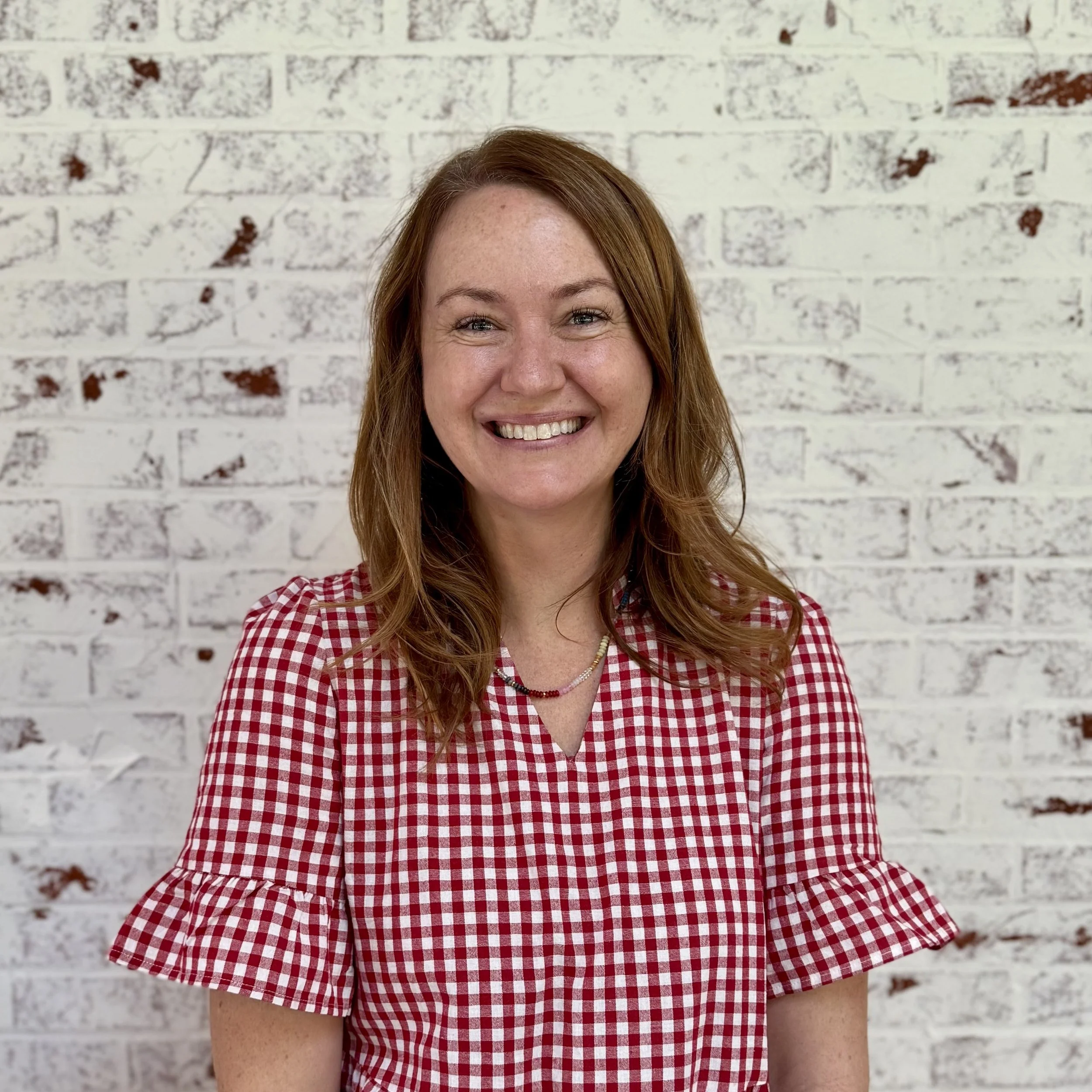 Smiling woman with red hair wearing a red and white checkered dress in front of a white brick wall.