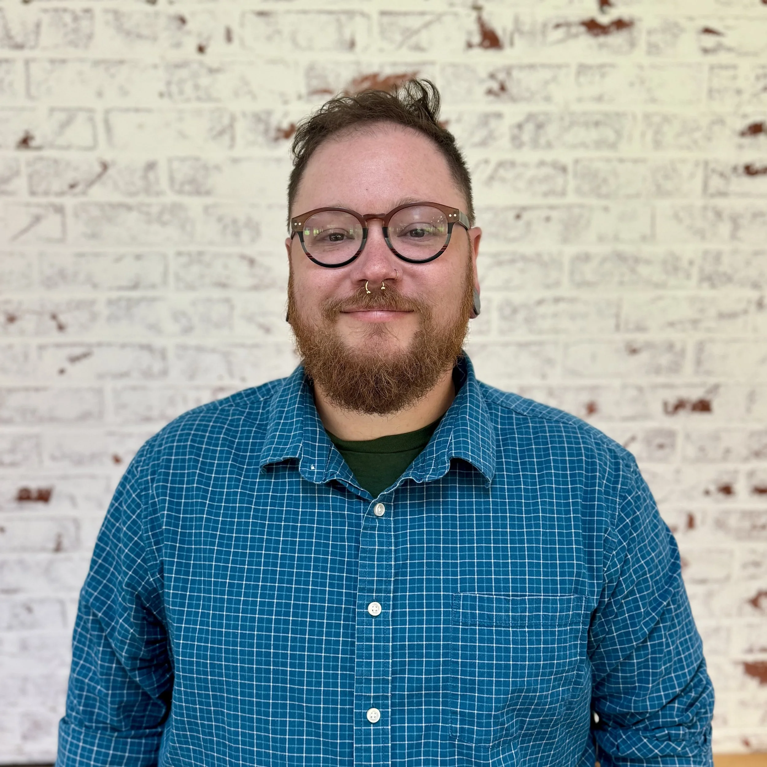 A man with short brown hair, glasses, and a beard, smiling and wearing a blue checkered shirt, standing against a white brick wall.
