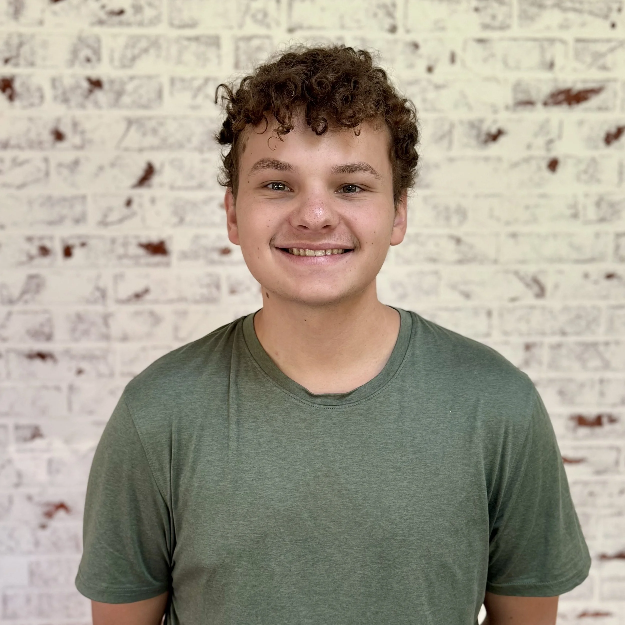 Smiling young man with curly brown hair wearing a green T-shirt standing in front of a white brick wall.