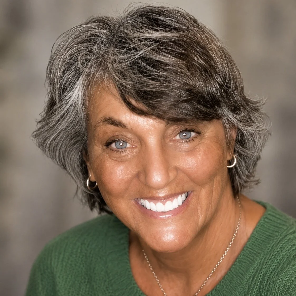 A smiling woman with short gray hair, blue eyes, and earrings, wearing a green top.