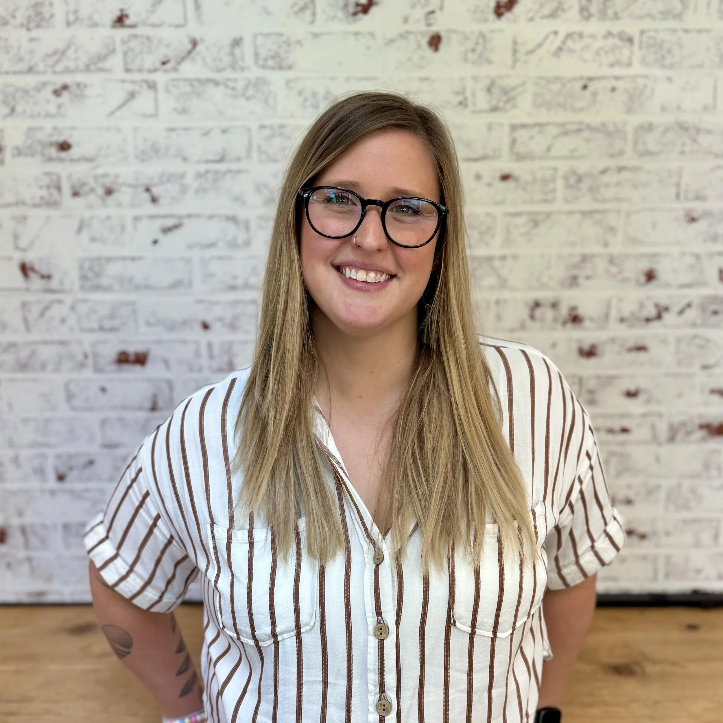 A woman with long blond hair, wearing glasses, smiling, standing in front of a white brick wall, dressed in a white shirt with brown stripes.