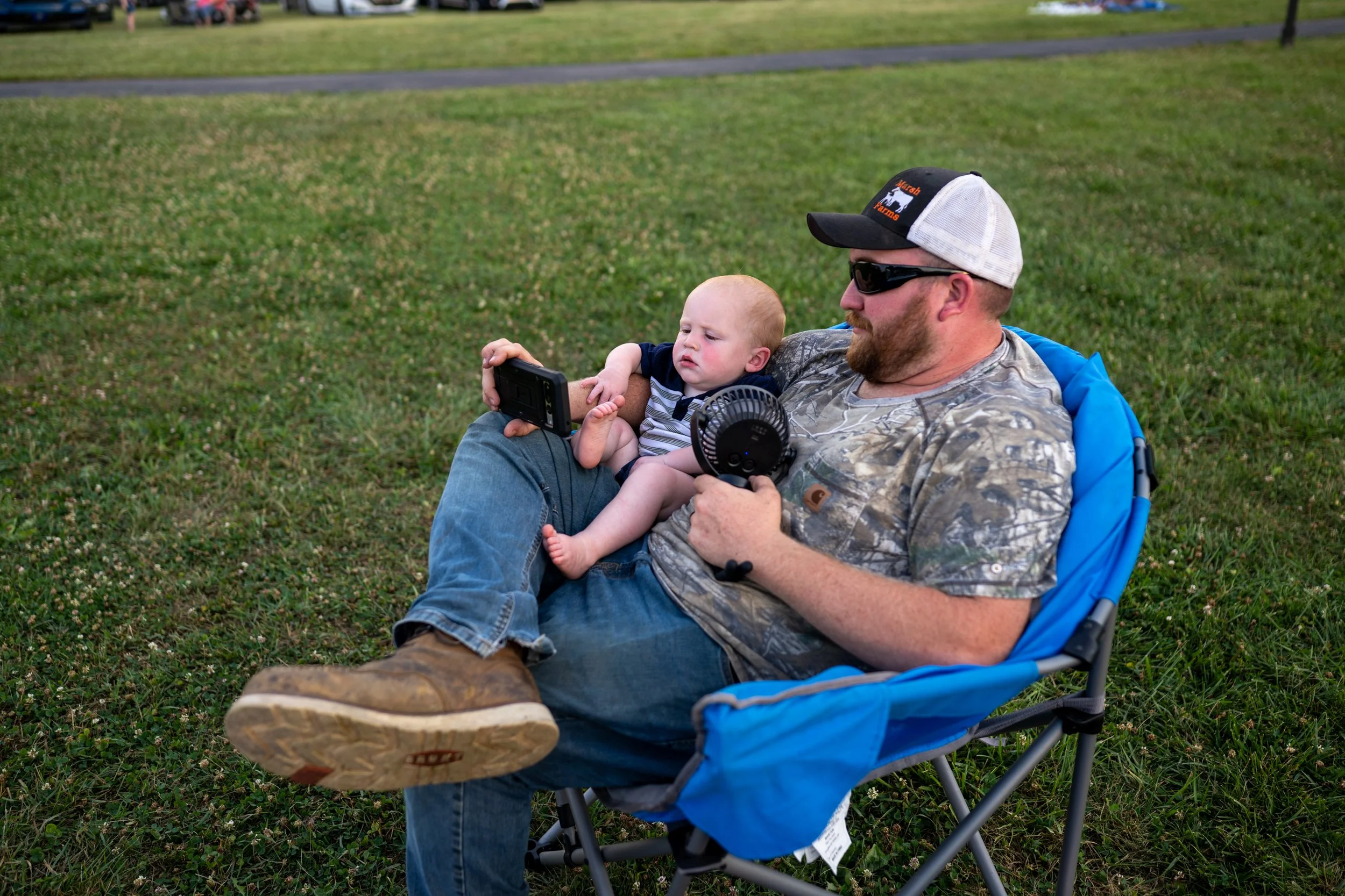 Adam Marsh, 34, watches a show and fans his son, James Marsh, 1, at the Rotary Club's Fourth of July celebration at Flat Run’s Veterans Park on Saturday, June 29, 2024. The temperatures were in the upper 90ºs. 