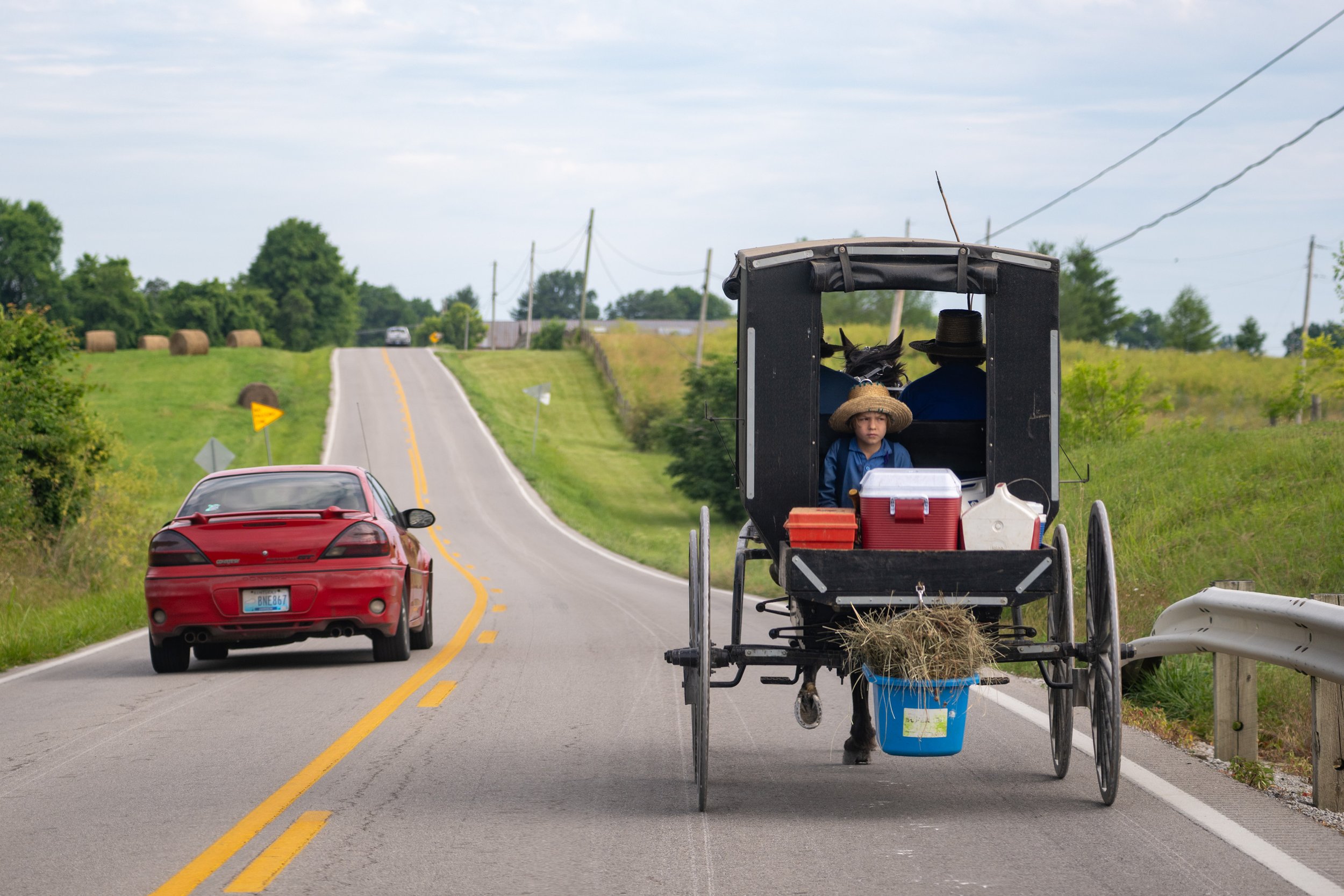 Amish members Mahlon Hostetler, John Gingerich, and Samuel Gingerich, 6, are passed by a car as they head to work by horse and buggy on Lafferty Pike Road near Berry, Kentucky on Friday, June 14, 2024. Part of Boyd's Station Project 306.36 on rural A