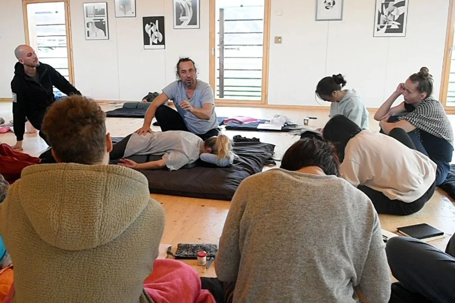 Groupe de personnes en atelier de méditation ou de relaxation, certains assis ou allongés sur des matelas, dans une pièce lumineuse avec des œuvres abstraites accrochées au mur.