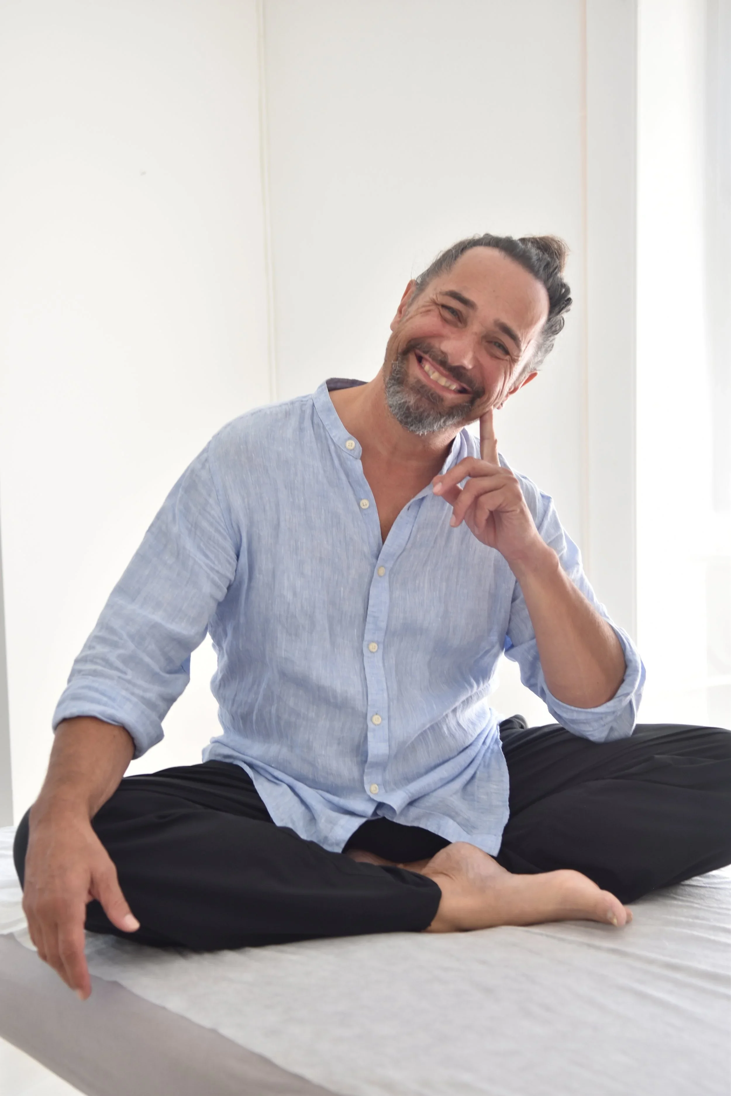 Jörg Schürpf., assis en position de méditation sur un matelas, portant un pantalon noir et une chemise bleue. Il touche doucement sa joue avec le doigt.