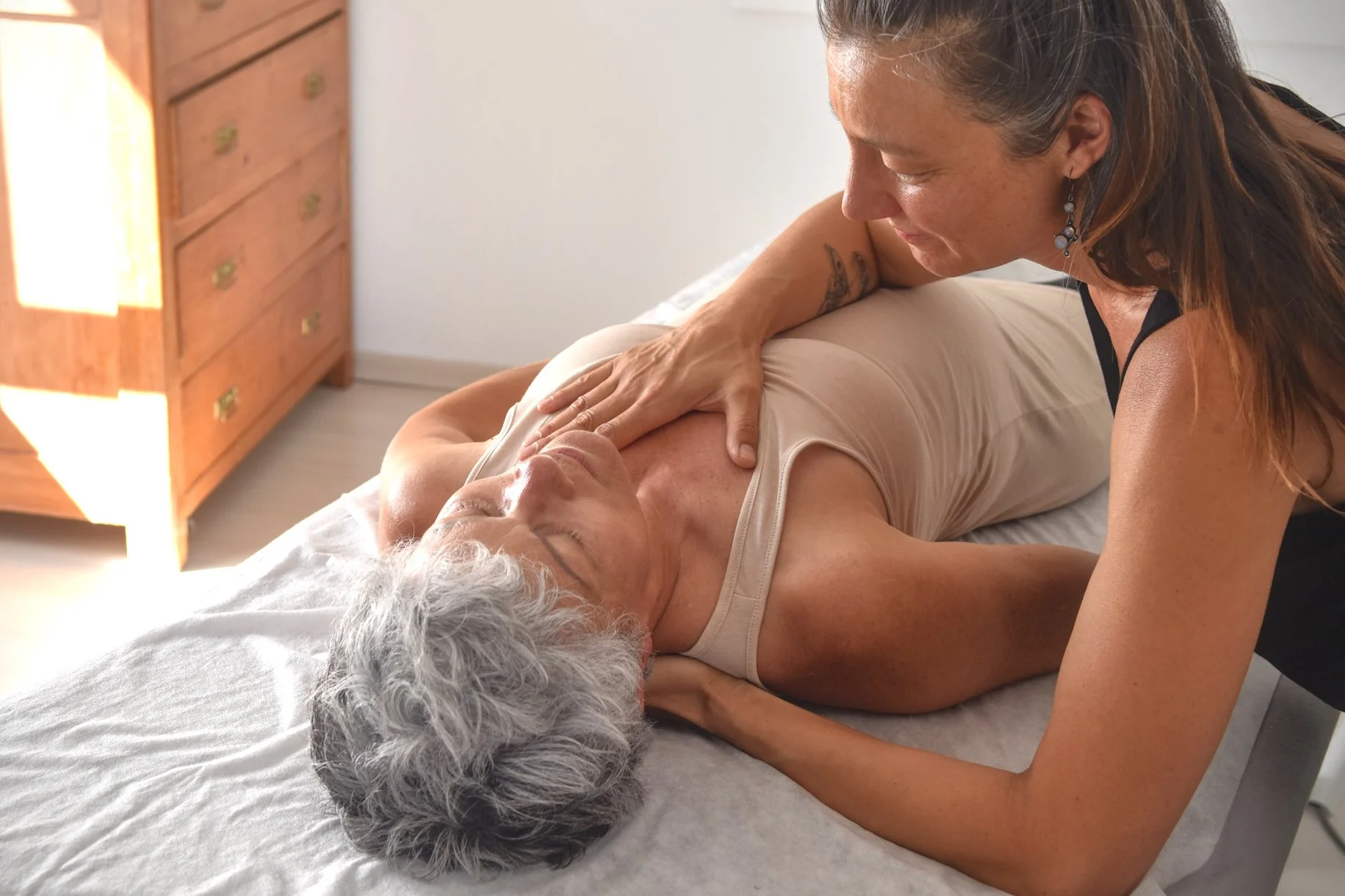 Une femme plus âgée reçoit un massage de la part d'une autre femme dans une pièce lumineuse, probablement une maison ou un salon de massage.