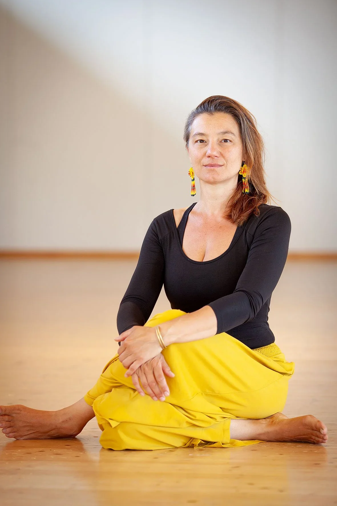 Muriel assise en position de yoga avec une jambe pliée et une main sur le genou, portant un haut noir, un pantalon jaune et des boucles d'oreilles colorées, dans une salle de yoga