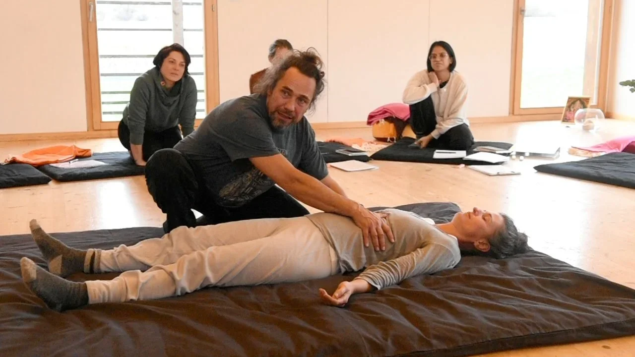 Une femme est allongée sur un matelas, une personne pratique une technique de massage ou de thérapie. Trois autres personnes sont présentes dans la pièce, probablement dans un cadre d'apprentissage ou de séance de groupe, avec des tapis et des documents sur le sol.