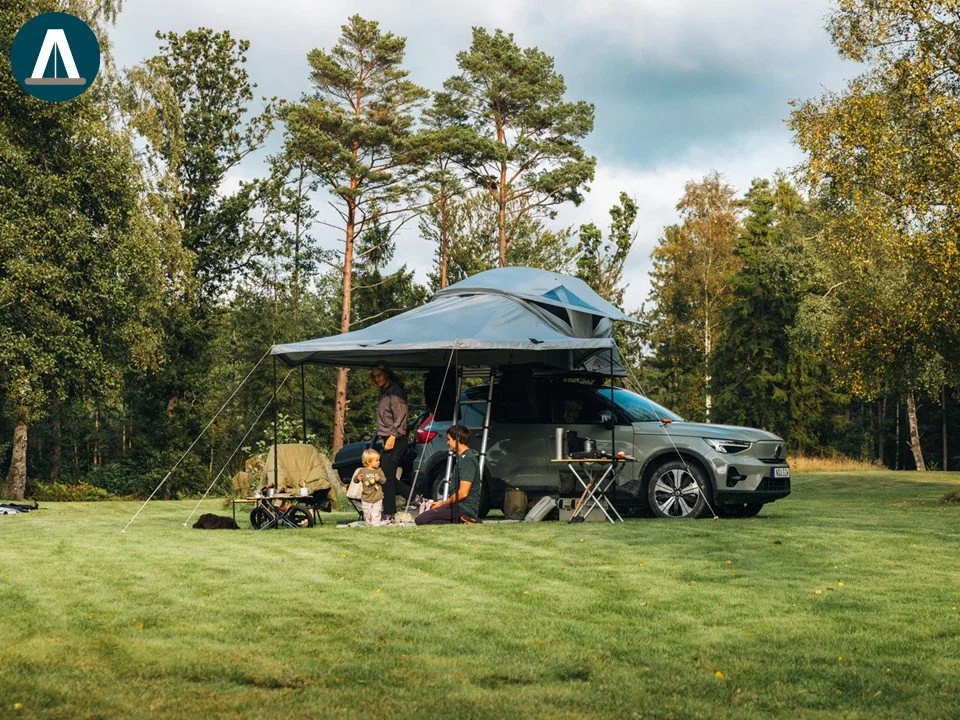 Mensen genieten van een campingweekend met een auto en een tent uitbouw, omgeven door bomen en gras, onder een bewolkte hemel.