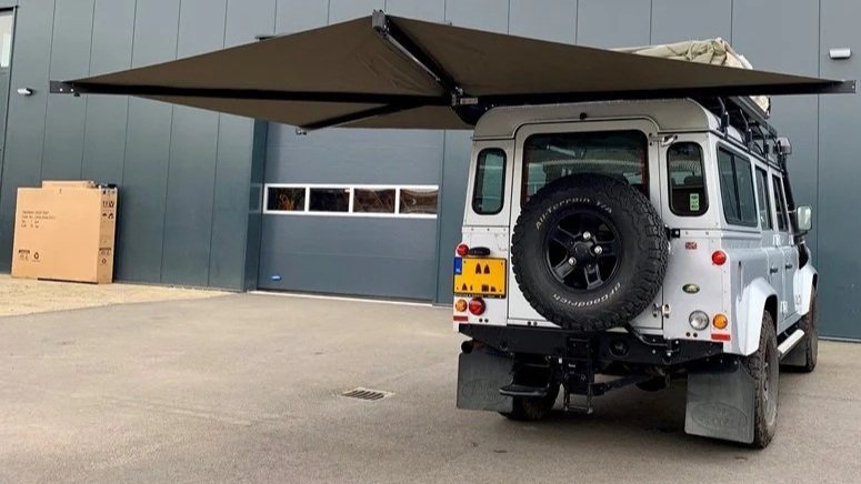 Een witte landrover met een daktentje en een uitklapbare luifel, geparkeerd op een parkeerplaats voor een grote, moderne loods met een garagepoort.