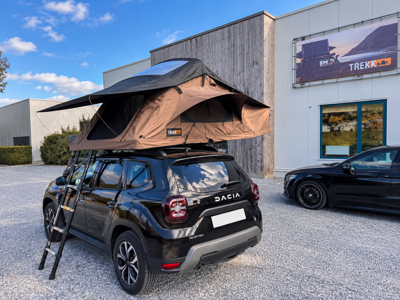 Een zwart Dacia Duster met een daktent en roltrap op de achterklep geparkeerd op een gravelparking voor een modern gebouw met borden en ramen, onder een blauwe hemel.