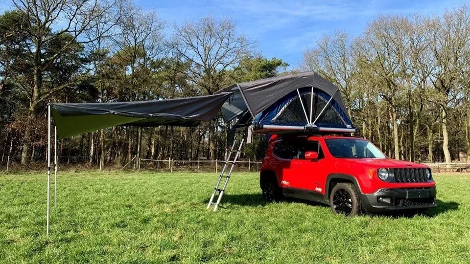 Een rode Jeep Renegade met een daktent en een uitklapbare luifel op een grasveld, omgeven door bomen.