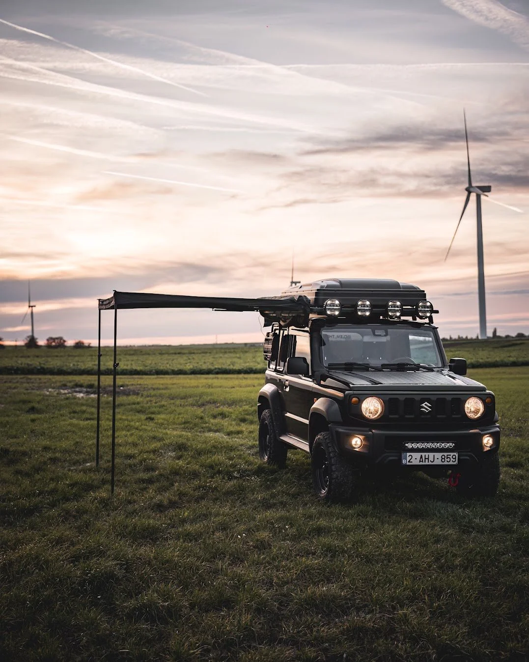 Een zwart Suzuki Jimny op een grasveld met windturbines op de achtergrond tijdens zonsondergang.