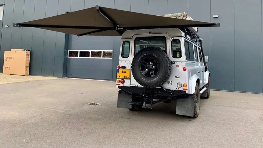 Een witte terreinwagen met een reservewiel aan de achterkant, een uitklapbare luifel en een dakdrager. De auto staat geparkeerd voor een grijze muur met een garagepoort.